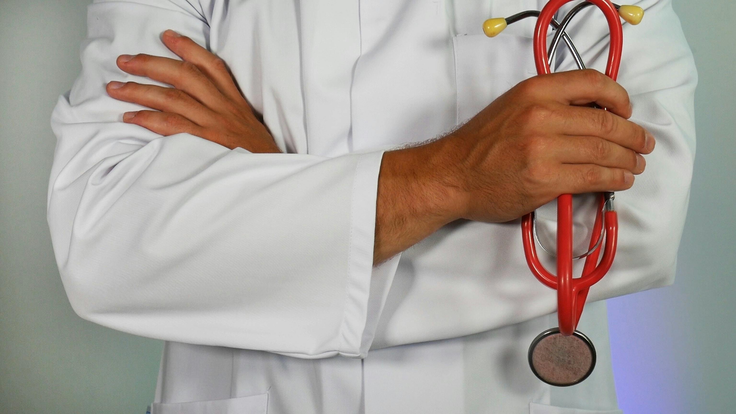Halbnah zeigt das Bild die verschränkten Arme eines Arztes in weißem Kittel mit rot-gelbem Stethoskop in einer Hand.