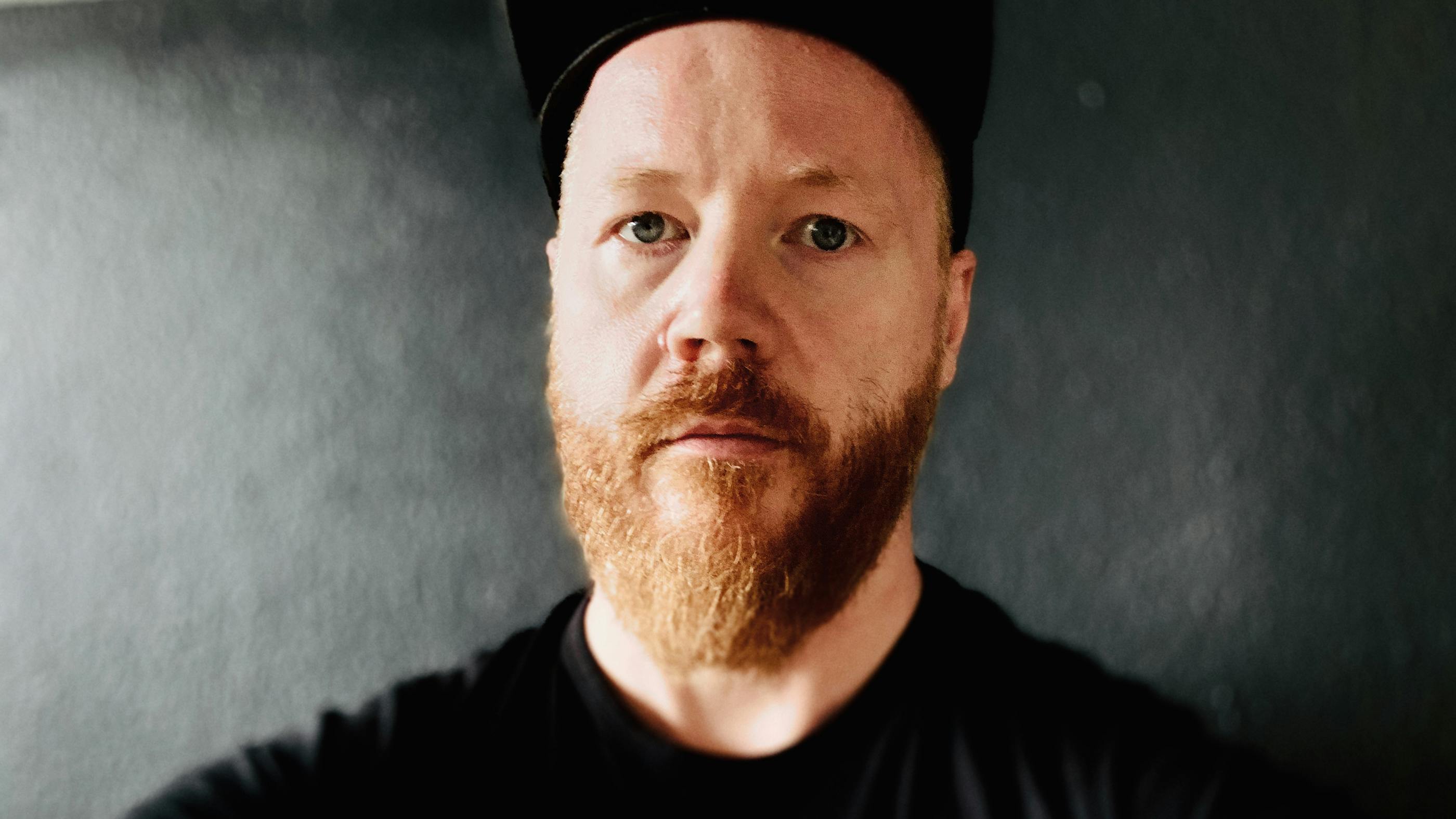 Ein Selfie von Autor Martin, im Hintergrund eine graue Wand. Martin hat blaue Augen, einen rötlichen Bart, trägt ein schwarzes T-Shirt und eine Basecap. 