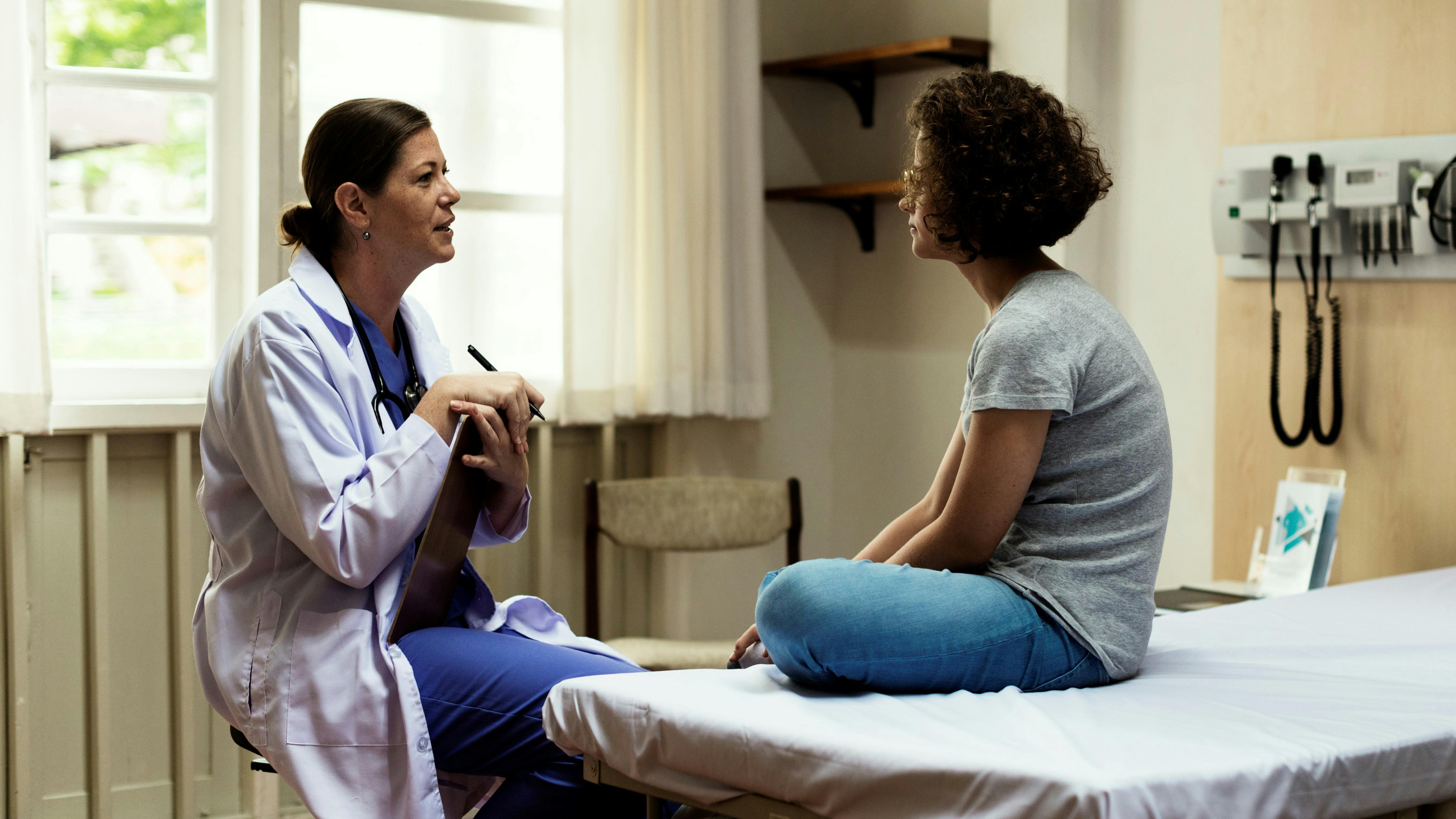 Eine Ärztin spricht im Krankenhaus mit einer Patientin, die im Schneidersitz auf dem Bett sitzt. 