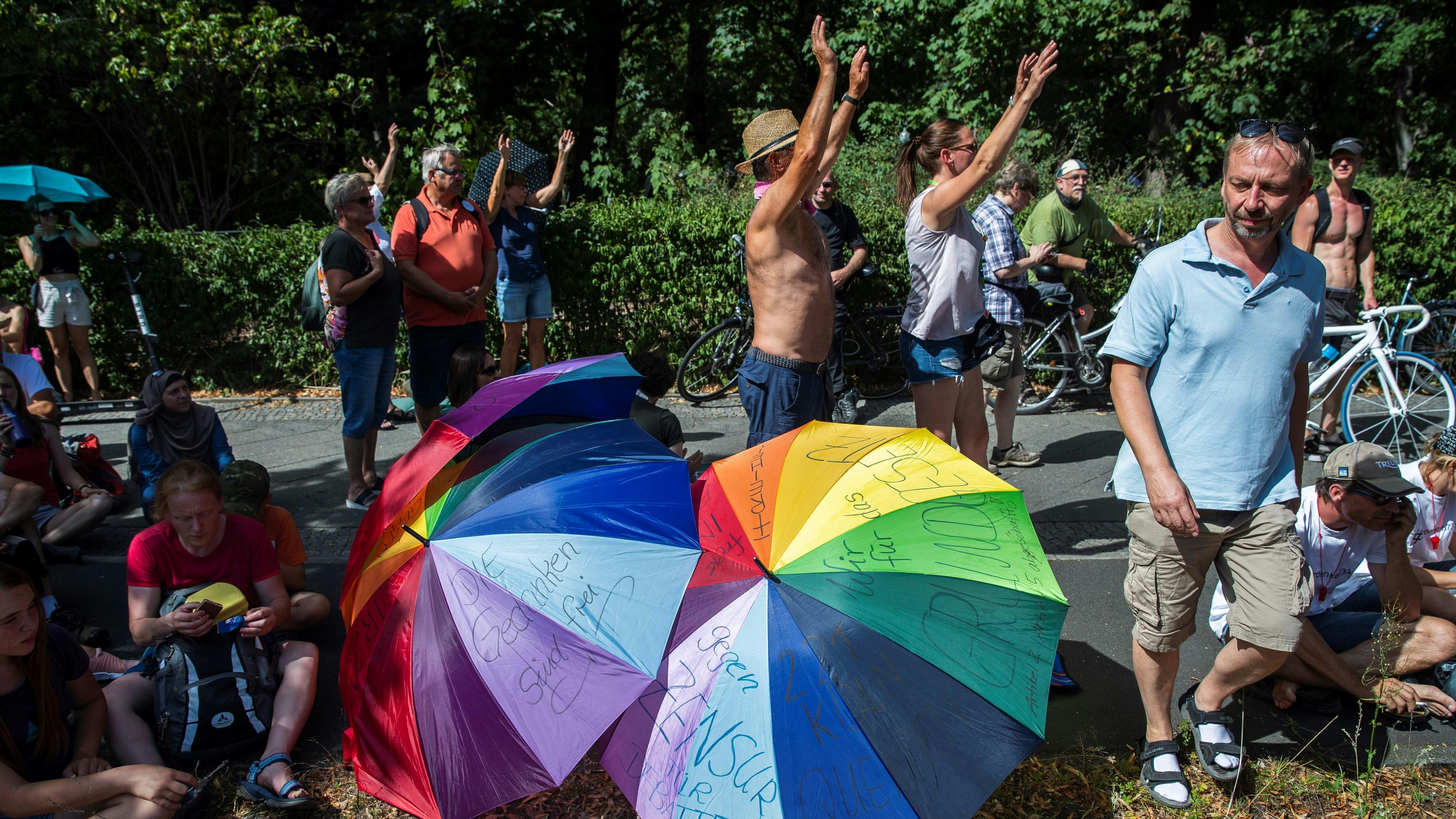 Corona-Demonstranten stehen mit erhobenen Händen neben regenbogenfarbenen Regenschirmen.