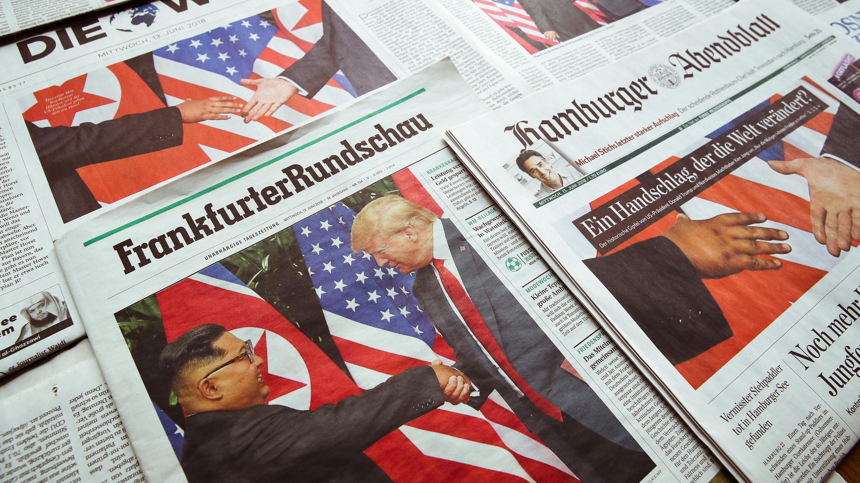 Wir sehen die Vorderseite von verscheidenen Zeitungen – auf allen ist ein Foto vom Trump und Kim jong un zu sehen, wie sie sich die Hand geben. 