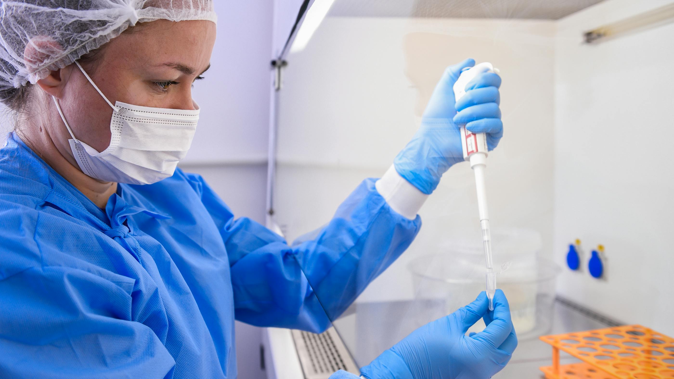 Wir sehen eine Wissenschaftlerin im Lab, die in voller blauer Montur und Mundschutz in einer Pipette eine Flüssigkeit zieht. 