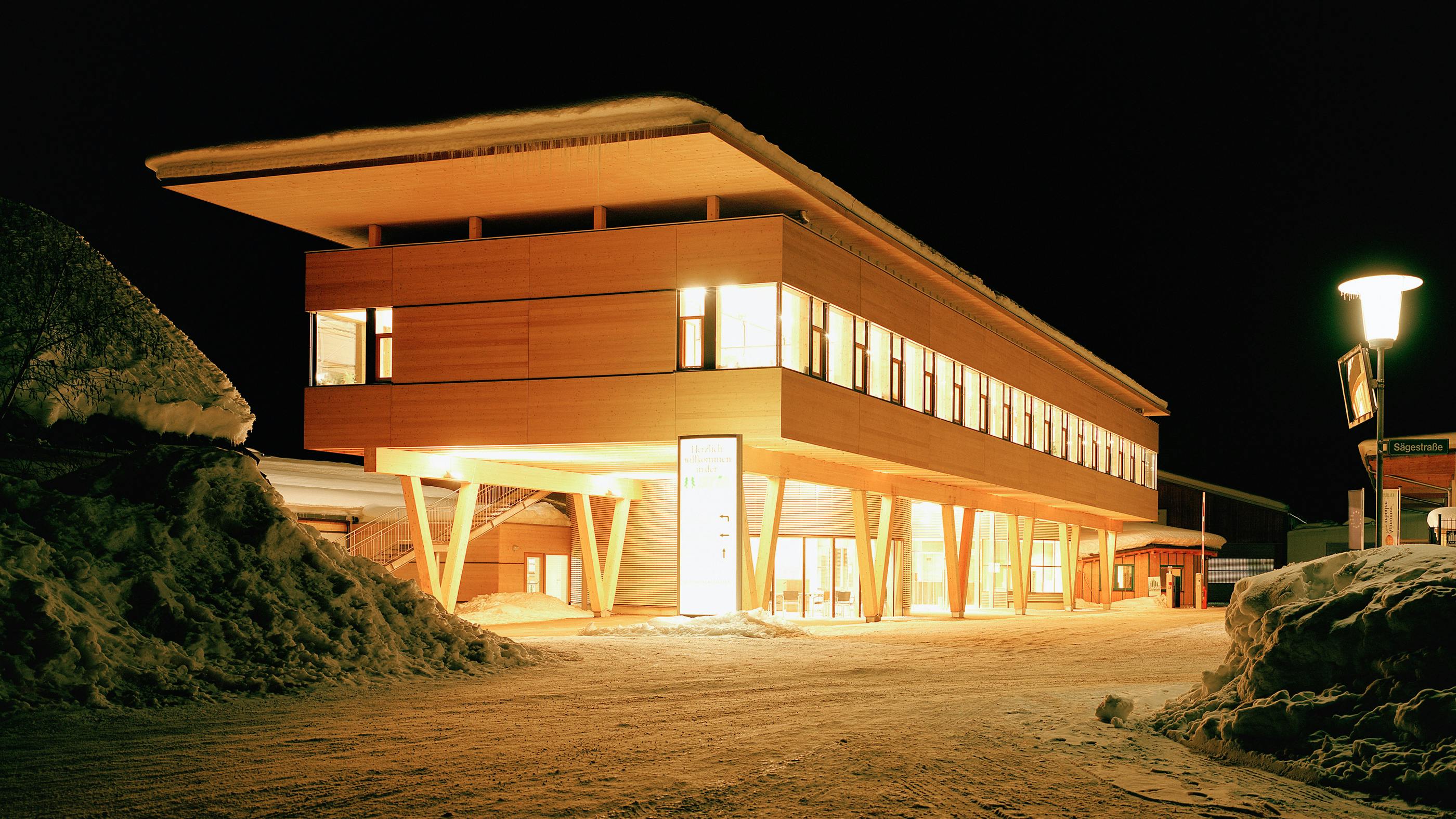 Wir sehen eine Nachtaufnahme eines längeren Gebäude, das komplett aus Holz besteht und von innen voll beleuchtet ist. 