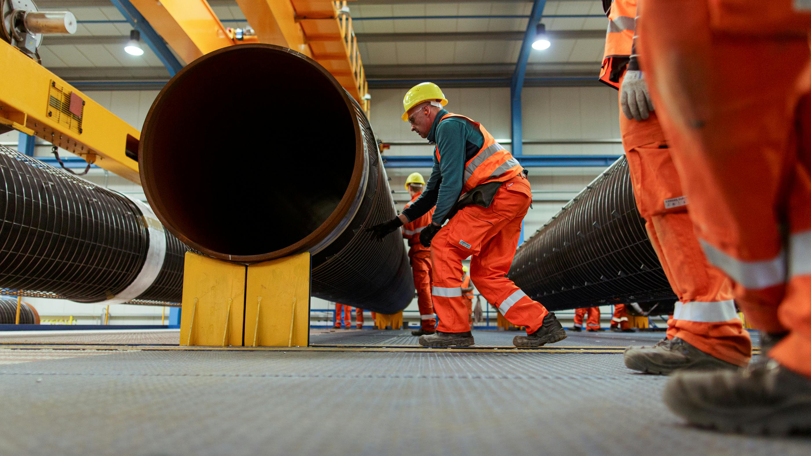 Wir sehen einen Arbeiter in oranener Arbeitskleidung, der unter ein großes Pipeline-Rohr fasst – ein paar Arbeiter schauen am rechten Rand zu.