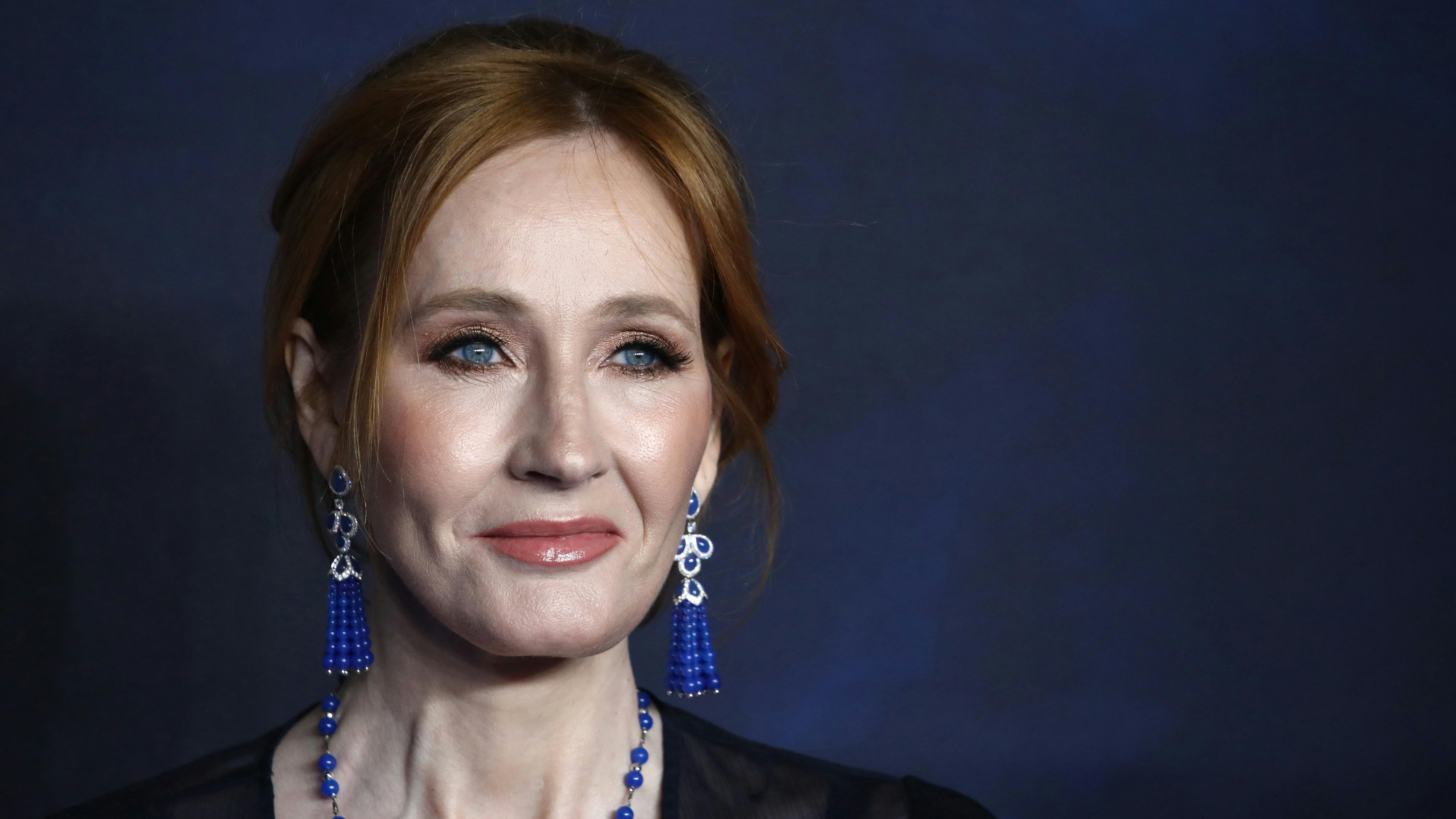 Wir sehen die Autorin Joanne K. Rowling im Portrait, lächelnd, vor einer dunkelblauen Wand. 