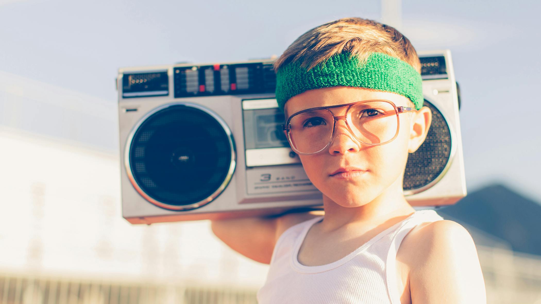 Ein kleiner Junge mit grünem Stirnband, Retro-Brille und armfreiem, geripptem Unterhemd posiert mit einem alten Radiorekorder auf der Schulter