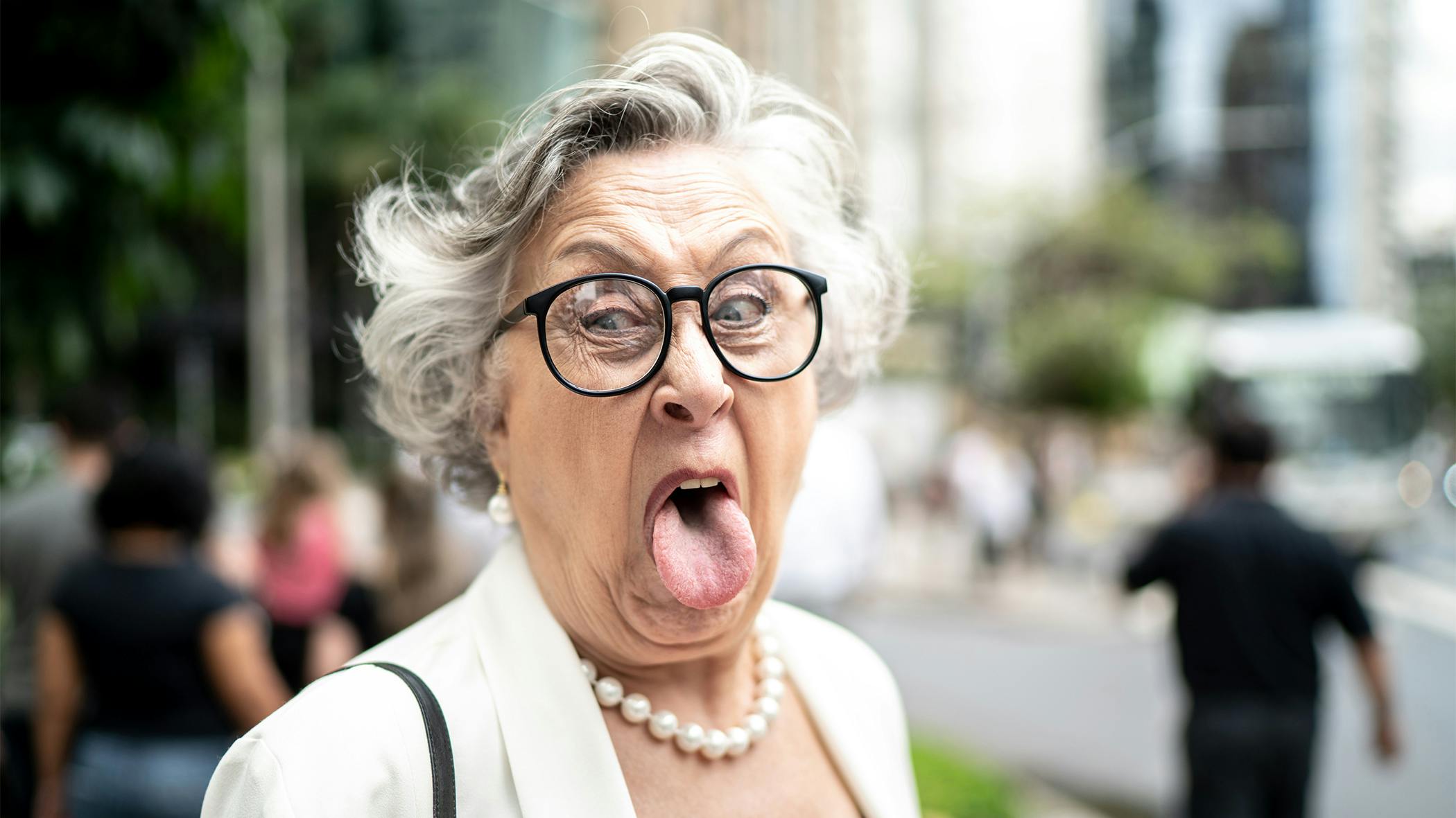 Eine ältere Dame mit weißen Haaren, Blazer, Perlenkette und Brille streckt mit verzerrtem Gesicht weit ihre Zunge heraus. 