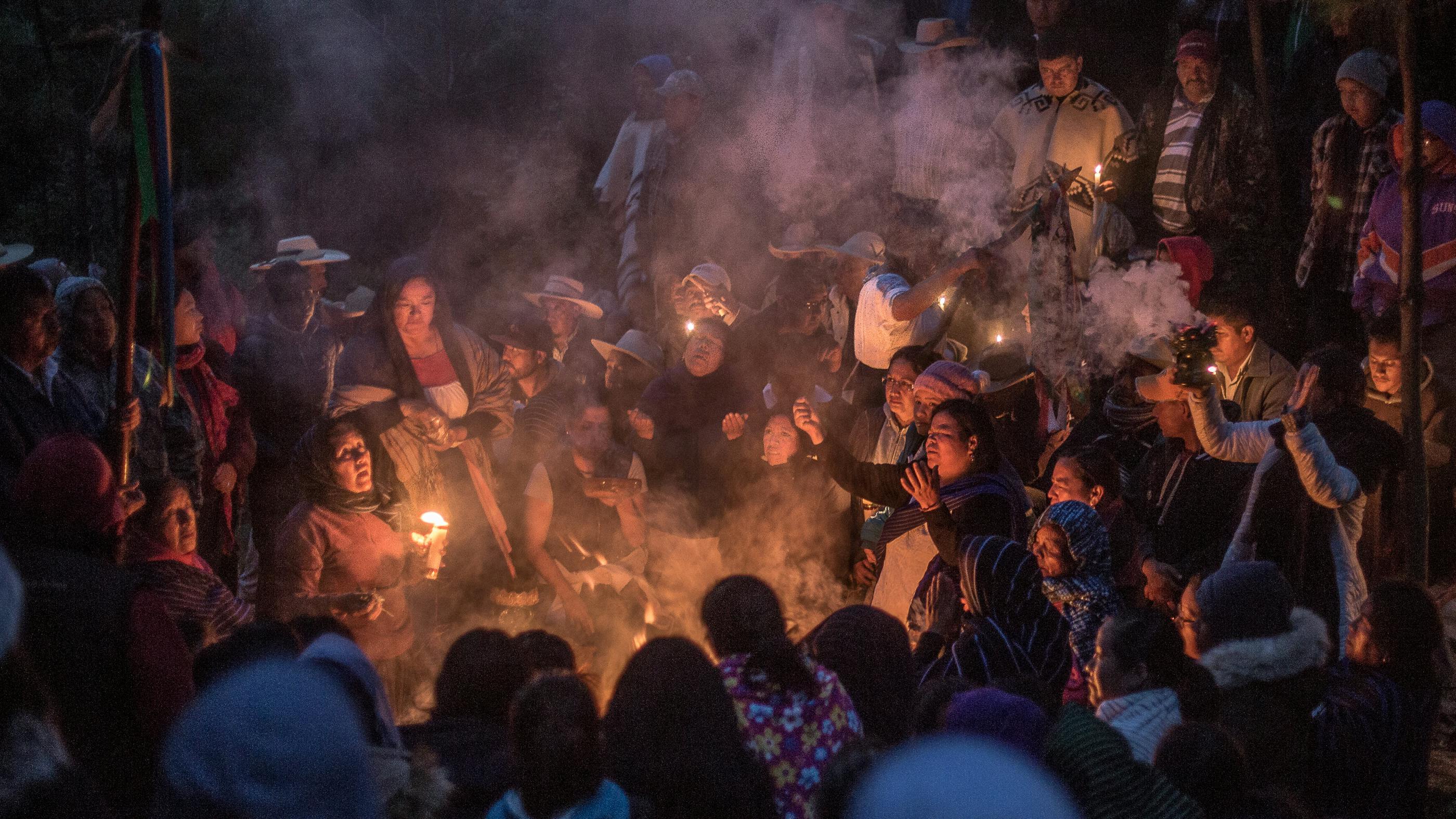 Eine größere Menschengruppe sitzt kreisförmig um ein Lagerfeuer, dahinter stehen weitere Menschen. Zwei Menschen am Feuer gestikulieren mit den Armen.