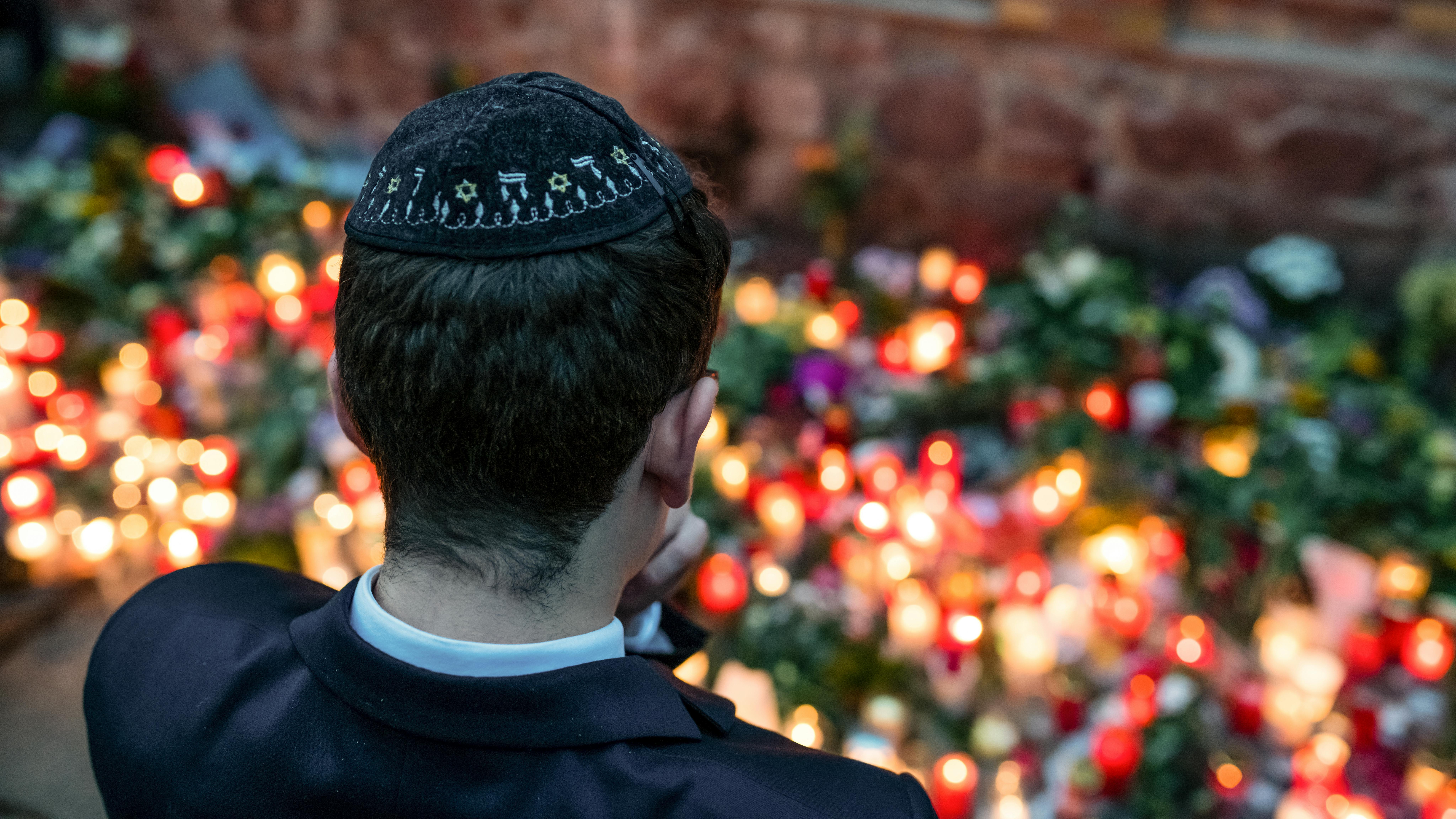Wir sehen einen jüdischen Jungen mit Kippa vor Blumen und Kerzen stehen. 