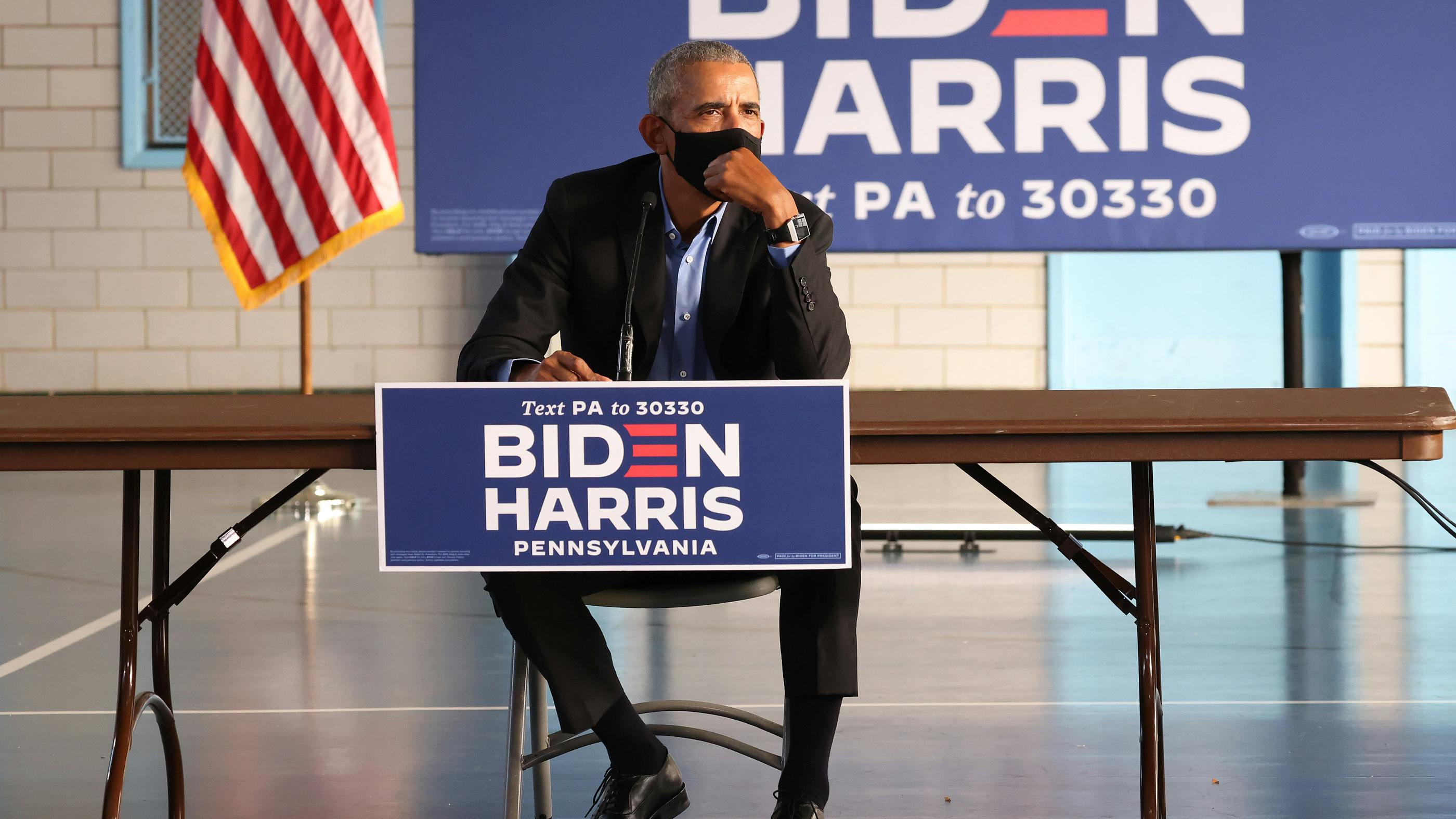 Ein nachdenklicher Barack Obama sietzt ein einem Tisch, dahinter BIDEN-HARRIS-Wahlplakate