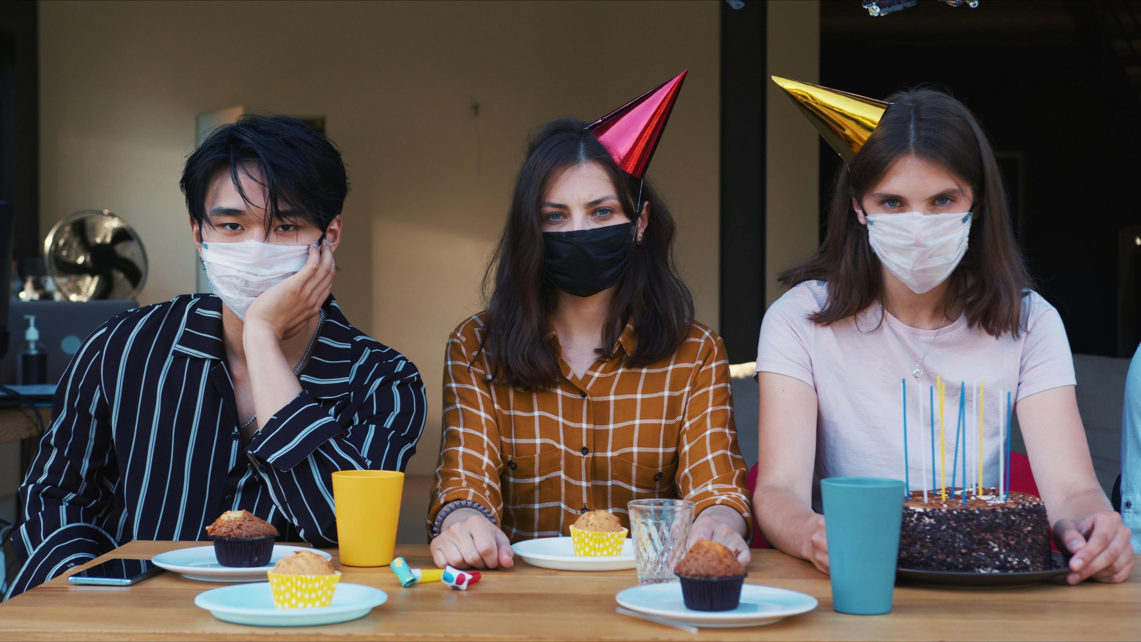 Drei Menschen sitzen mit Alltagsmasken an einem gedeckten Geburtstagstisch mit Kuchen. Sie sehen erschöpft und genervt aus.