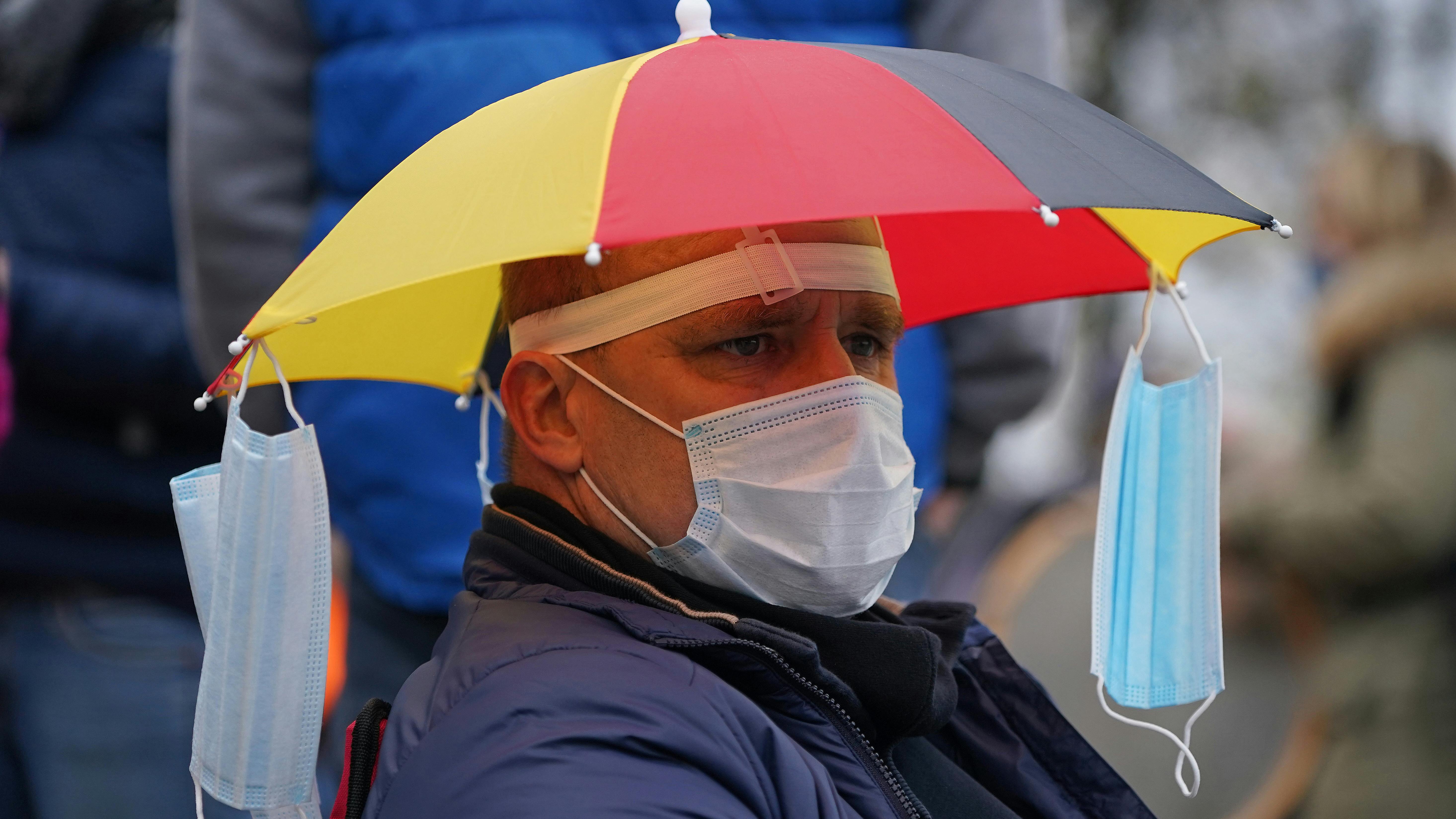 Ein Mann trägt einen Regenschirmhut in Deutschlandfarben, an dem Masken hängen. 