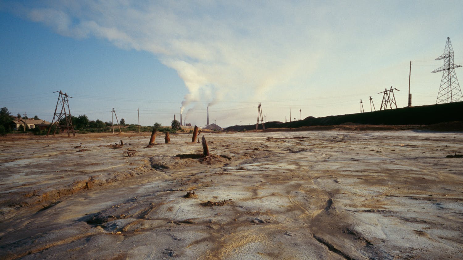 Ein erodierter Boden, einer Mondlandschaft gleich, mit stark rauchendem Schornstein im Hintergrund. Umweltverschmutzung in der Provinz Tscheljabinsk. Der Boden ist durch hohe Konzentrationen von Blei, Arsen, Nickel, Kobalt, Kadmium vergiftet.