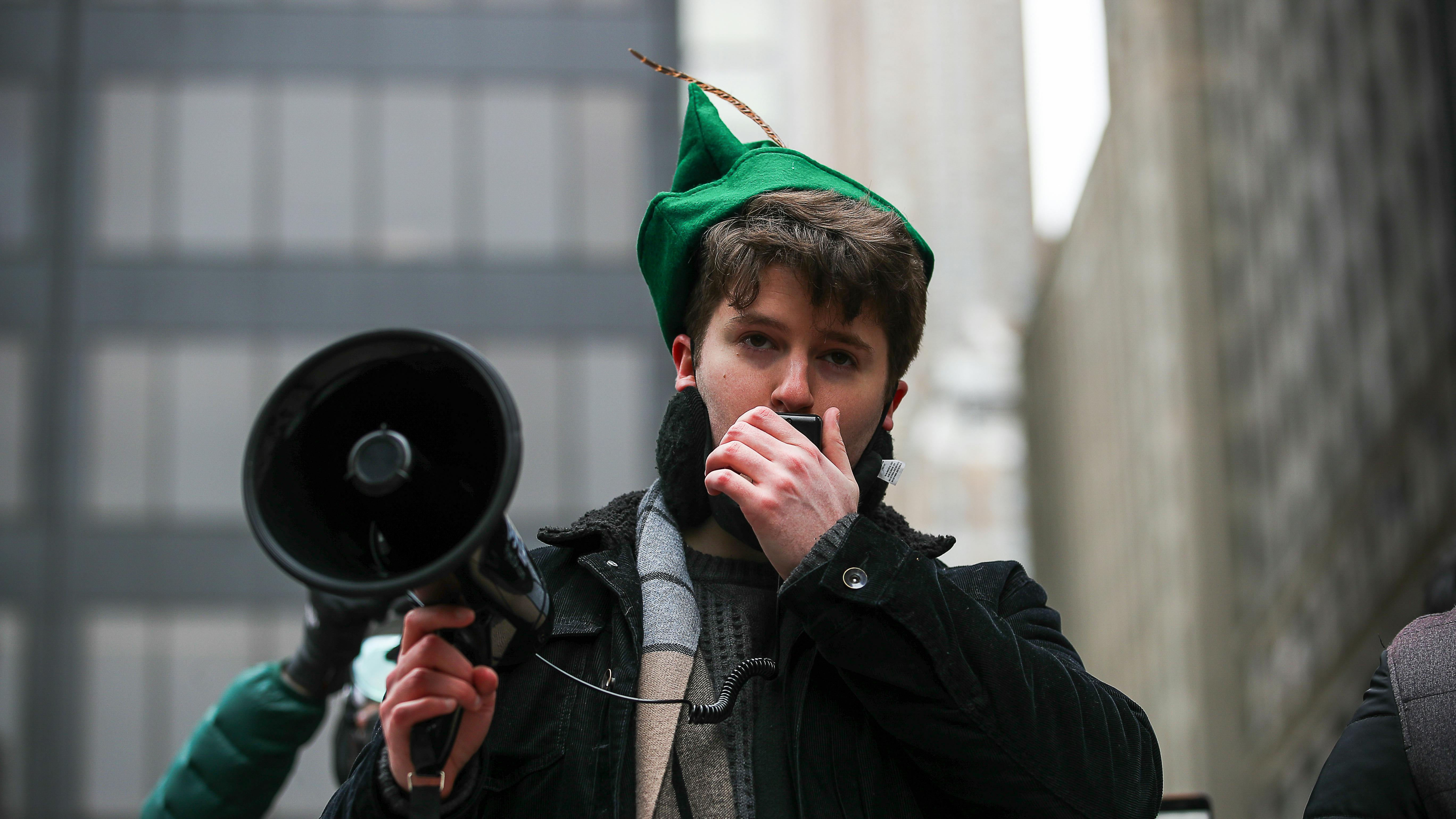 Ein jung-wirkender Mann mit altertümlicher Kappe spricht in ein Megaphone. 