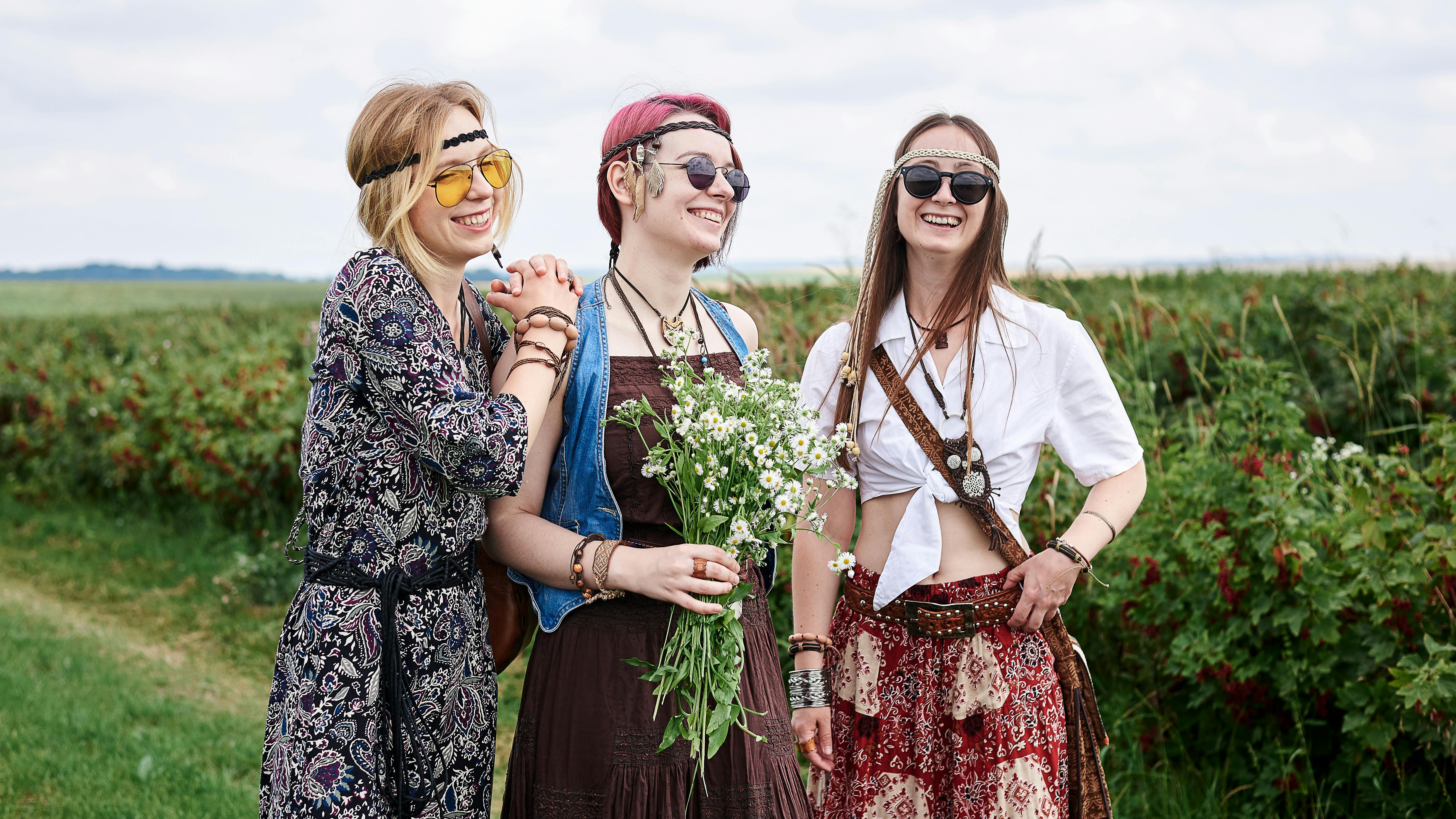 Drei junge Hippie-Frauen, die Kleidung im Boho-Stil tragen, stehen auf einem grünen Johannisbeerfeld und halten einen sommerlichen Kamillenstrauß in der Hand. 