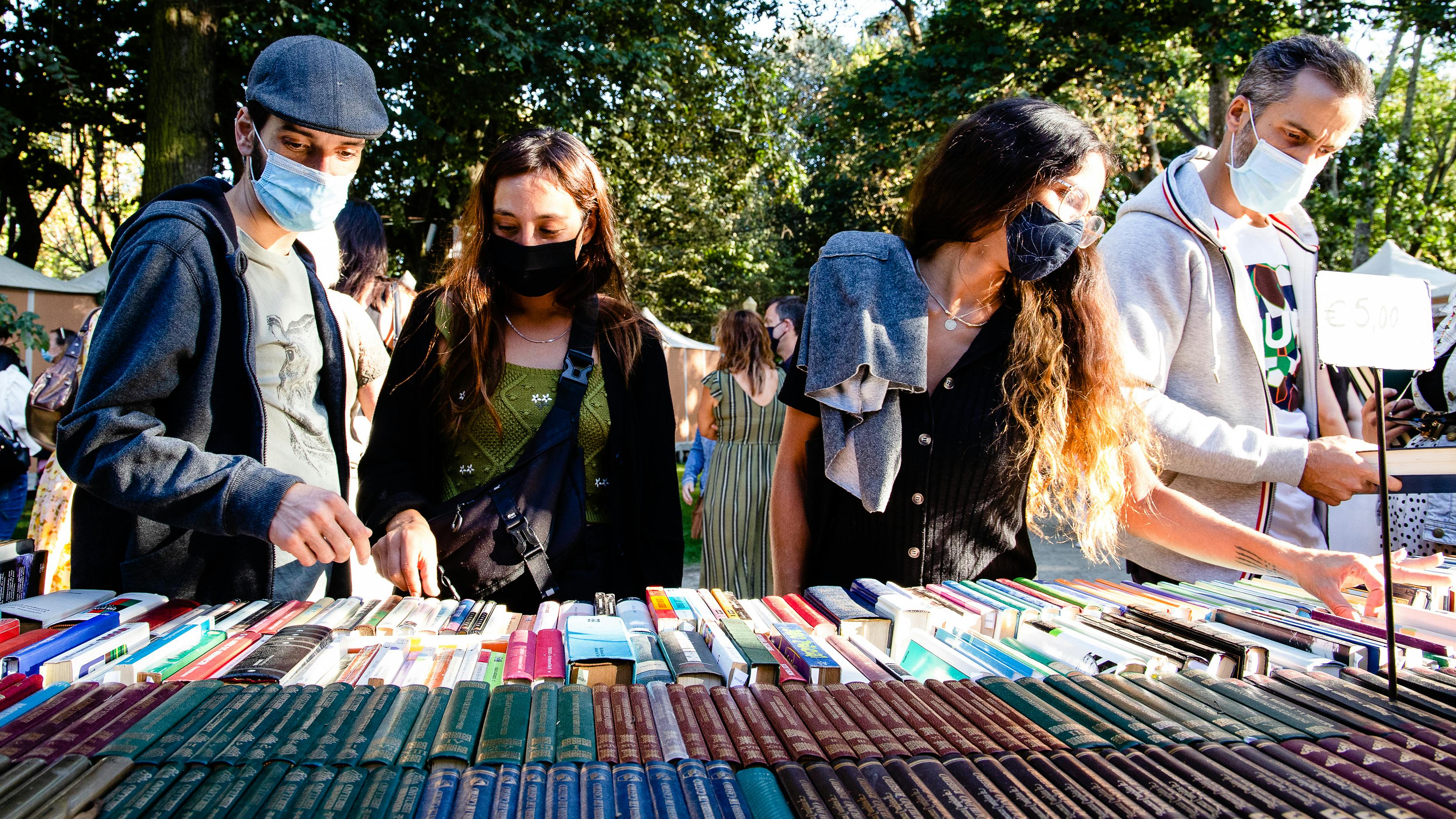 Eine Gruppe von vier Menschen mit Mundnasenschutz schauen sich Bücher in einer frühlingshaften Freiluft-Umgebung an.