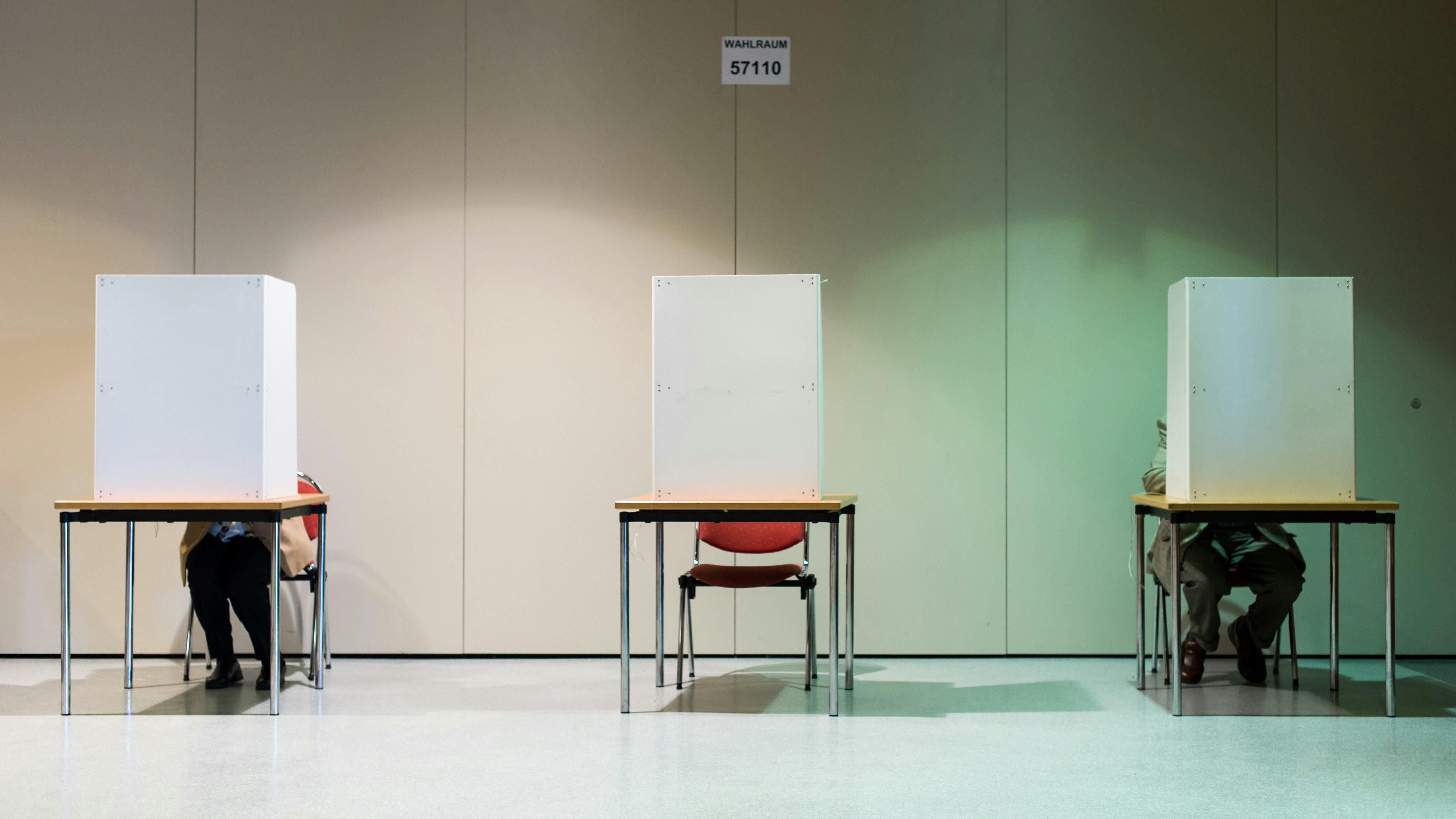 Drei Wahlkabinen stehen vor einer Wand mit Abstand im leichten Zwielicht aufgebaut, eine Person scheint in der äußerst linken zu sitzen. 