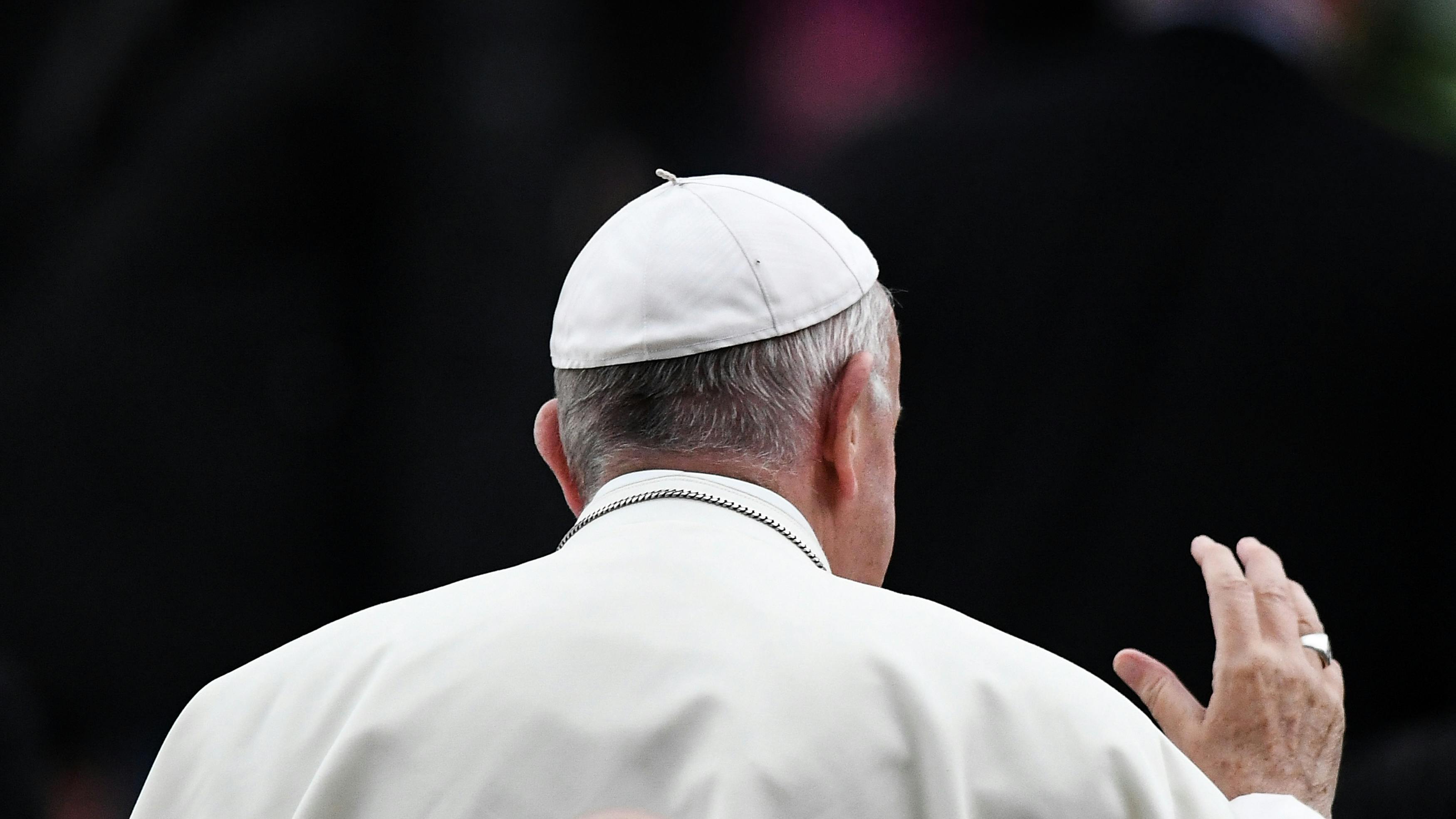 Wir sehen den Rücken des Papstes, durch seine Kopfbedeckung in Form kleinen Rundkappe, und sein weißes Gewand klar erkennbar. Er hebt die Hand zum große einer nicht sichtbaren Menge oder Person.