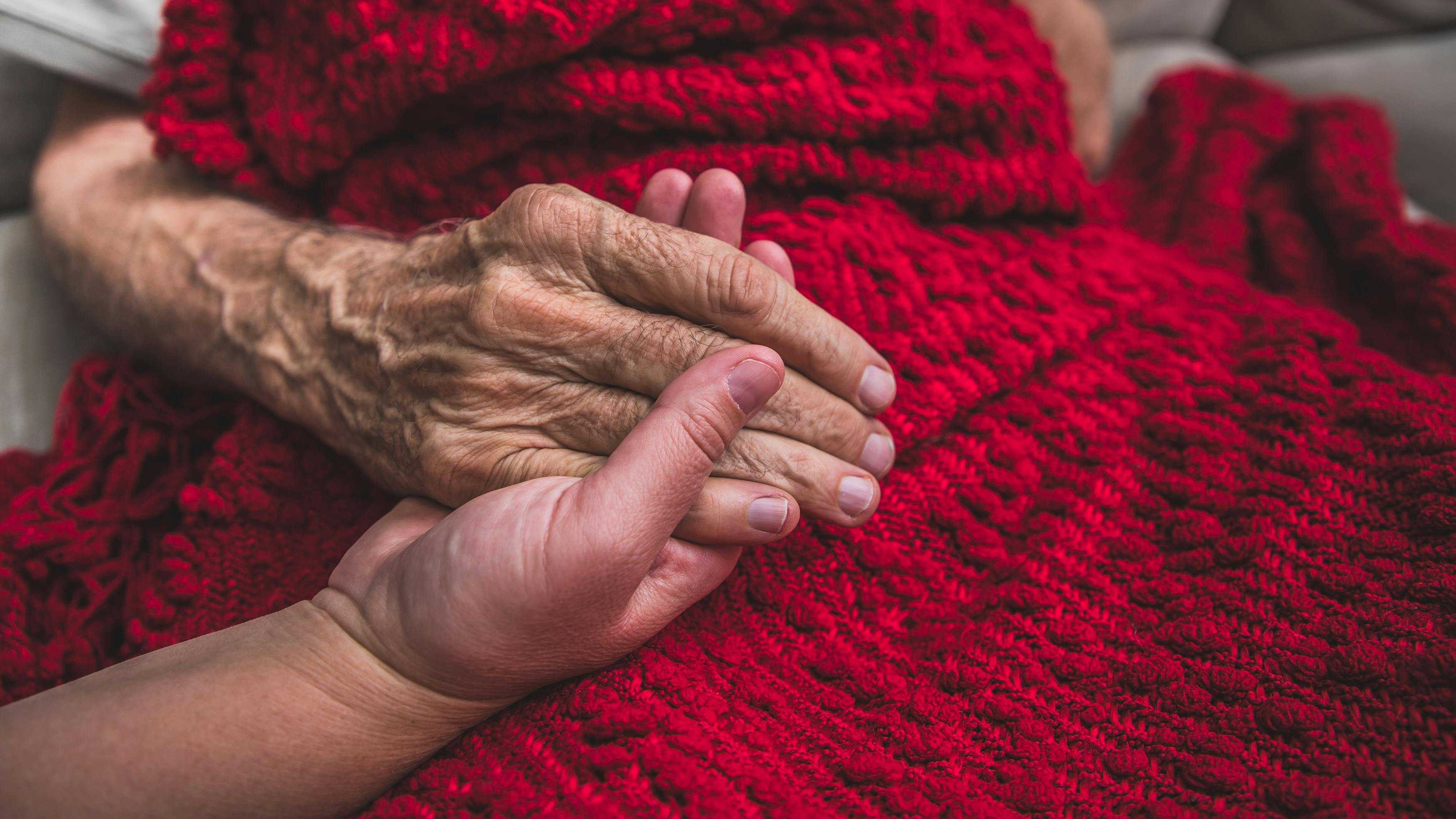 Eine stark gealterte und verwitterte Hand liegt auf einer roten Decke, von der Position her können wir erkennen das die Person sitzt. Die Hand wird von einer jünger aussehenden sanft gehalten.