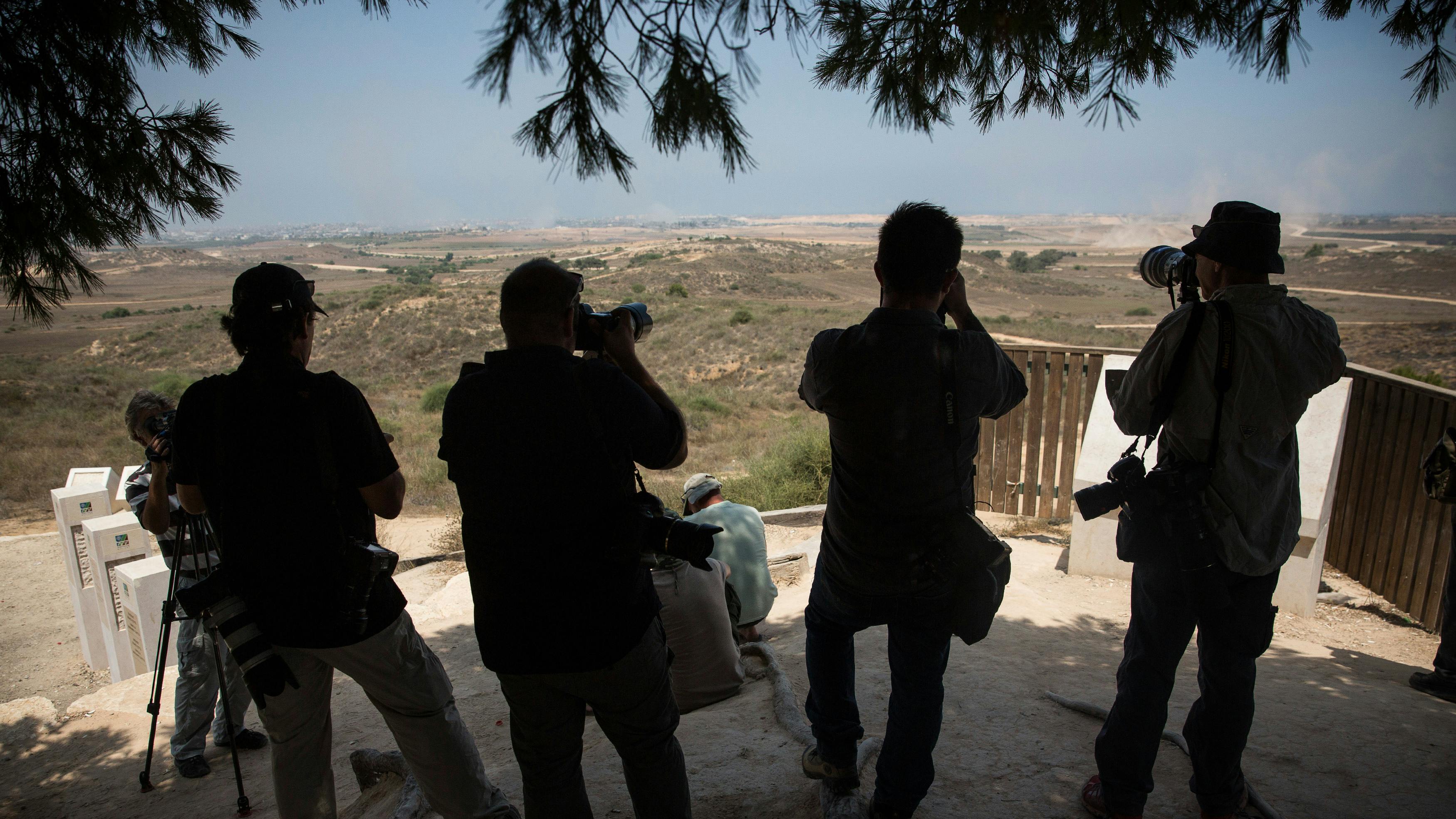 Im Vordergrund stehet eine Gruppe Fotograf*innen im Schatten eines Baumes, sie haben Ultratele-Objektive auf den Mittelgrund gerichtet. Dort sehen wir eine Staubfahne welche der Bildbeschreibung folgenden von, auf Gaza vorrückenden, Militärkonvois stammt. 