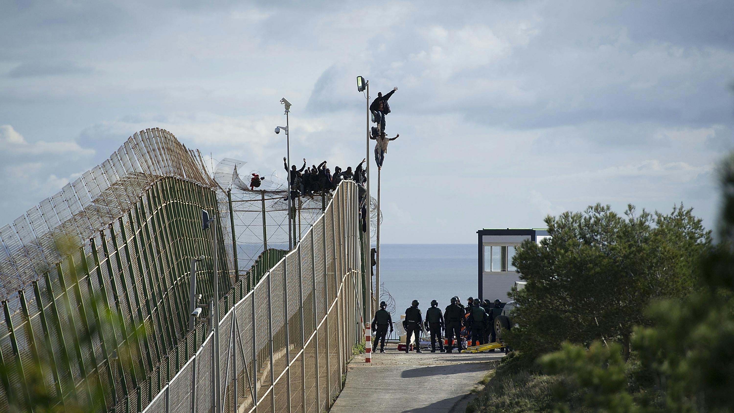 Eine Gruppe Migrant*innen überquert den Grenzzaun bei Mellila. Wir sehen den breiten  Zaun und eine Person hält ihre Hand hoch auf dem Zaun hängend. 