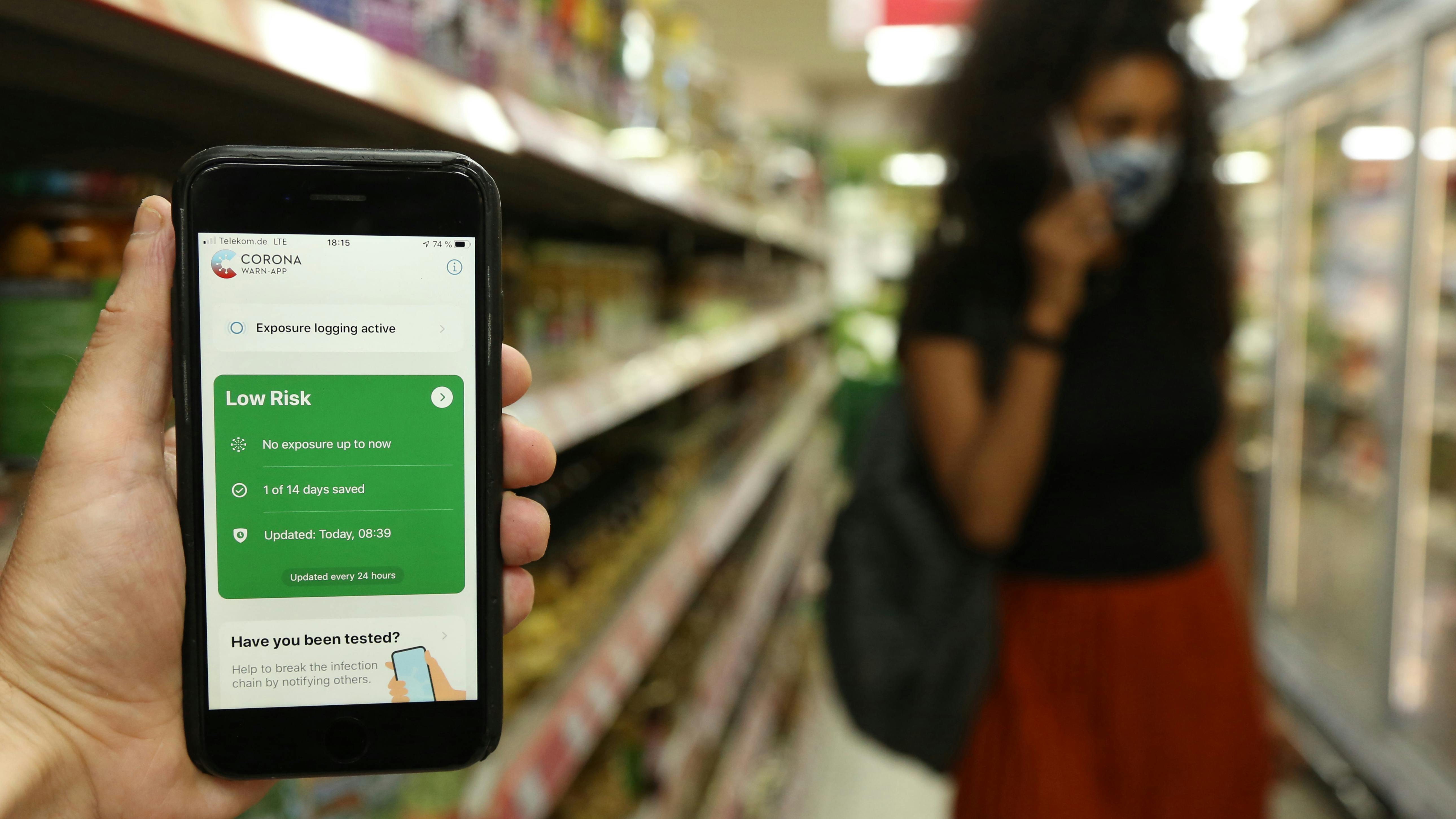 Ein Smartphone mit der Corona Warn App aufgerufen vor dem verschwommenen Hintergrund eines Supermarktgangs.