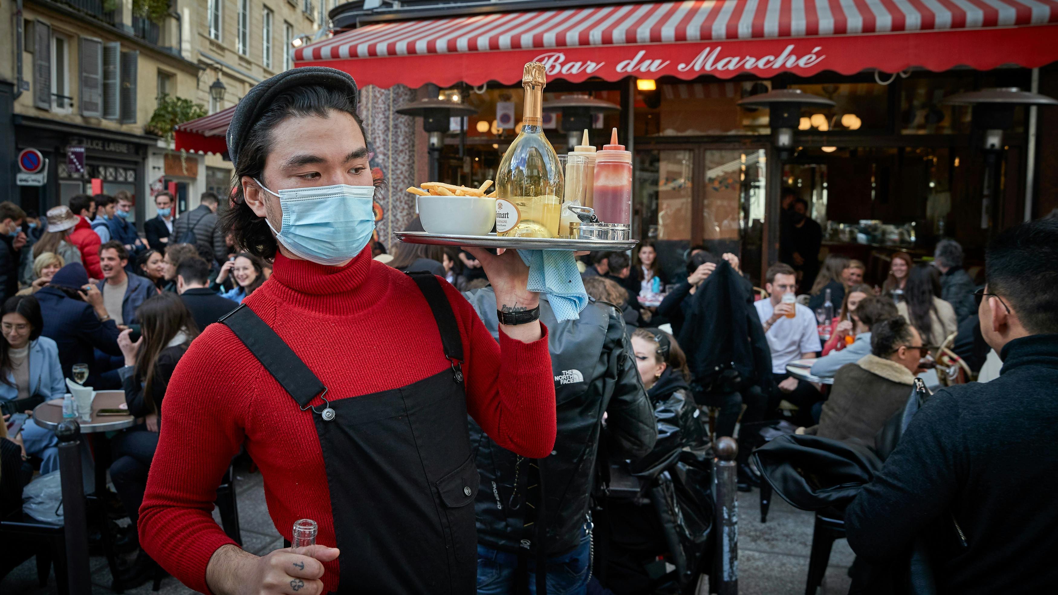 Ein Kellner mit Maske trägt ein volles Tablett durch den überfüllten Außenbereich eines Restaurants. 