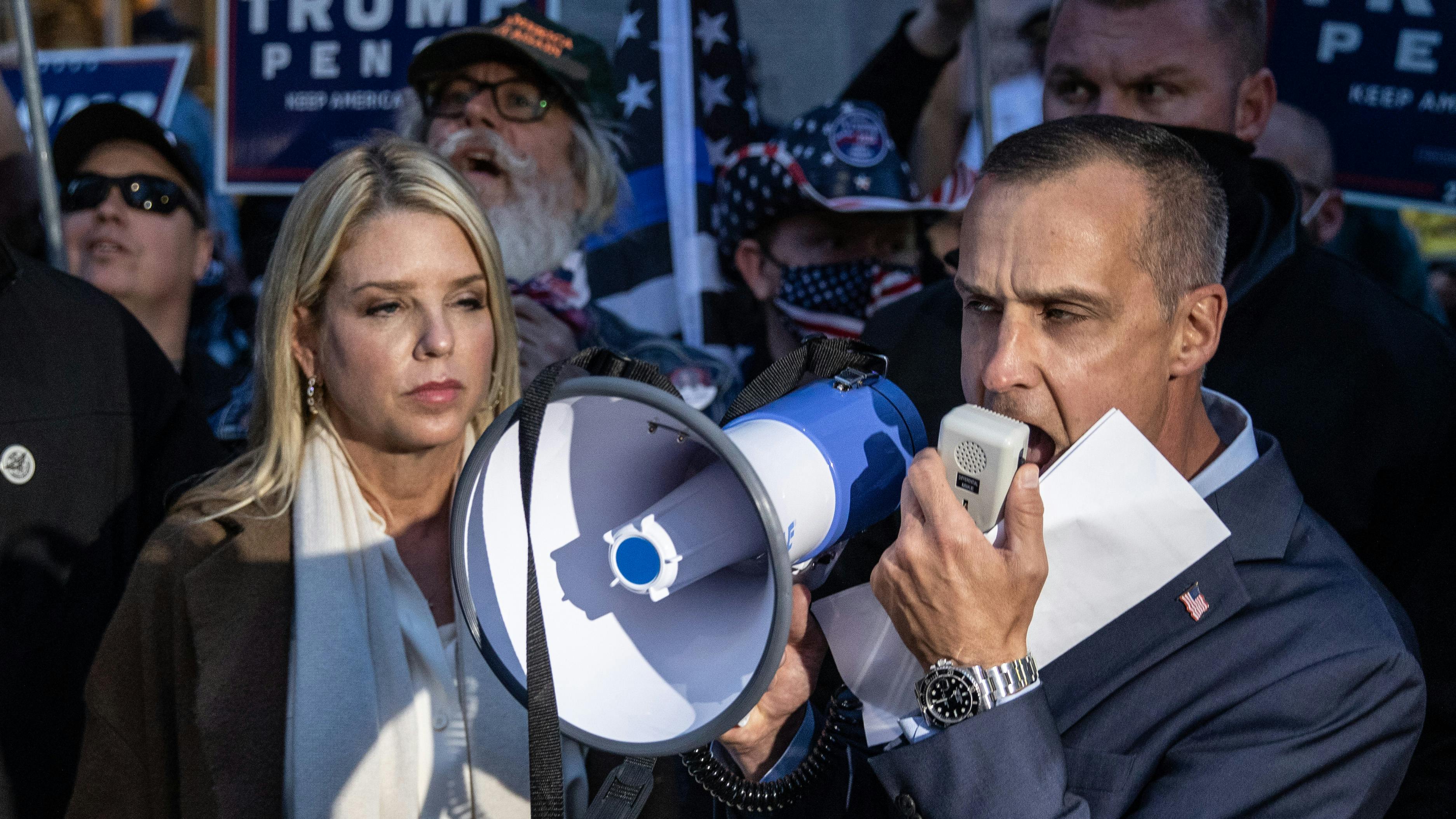 Ein Mann sprcht in ein Megaphon mit erzürntem Gesichtsausdruck. Neben ihm steht eine Frau welche entmutig und erschöpft scheint. Im Hintergrund sehen wir Pro-Trump Wahlposter welchen von AnhängerInnen gehalten werden.