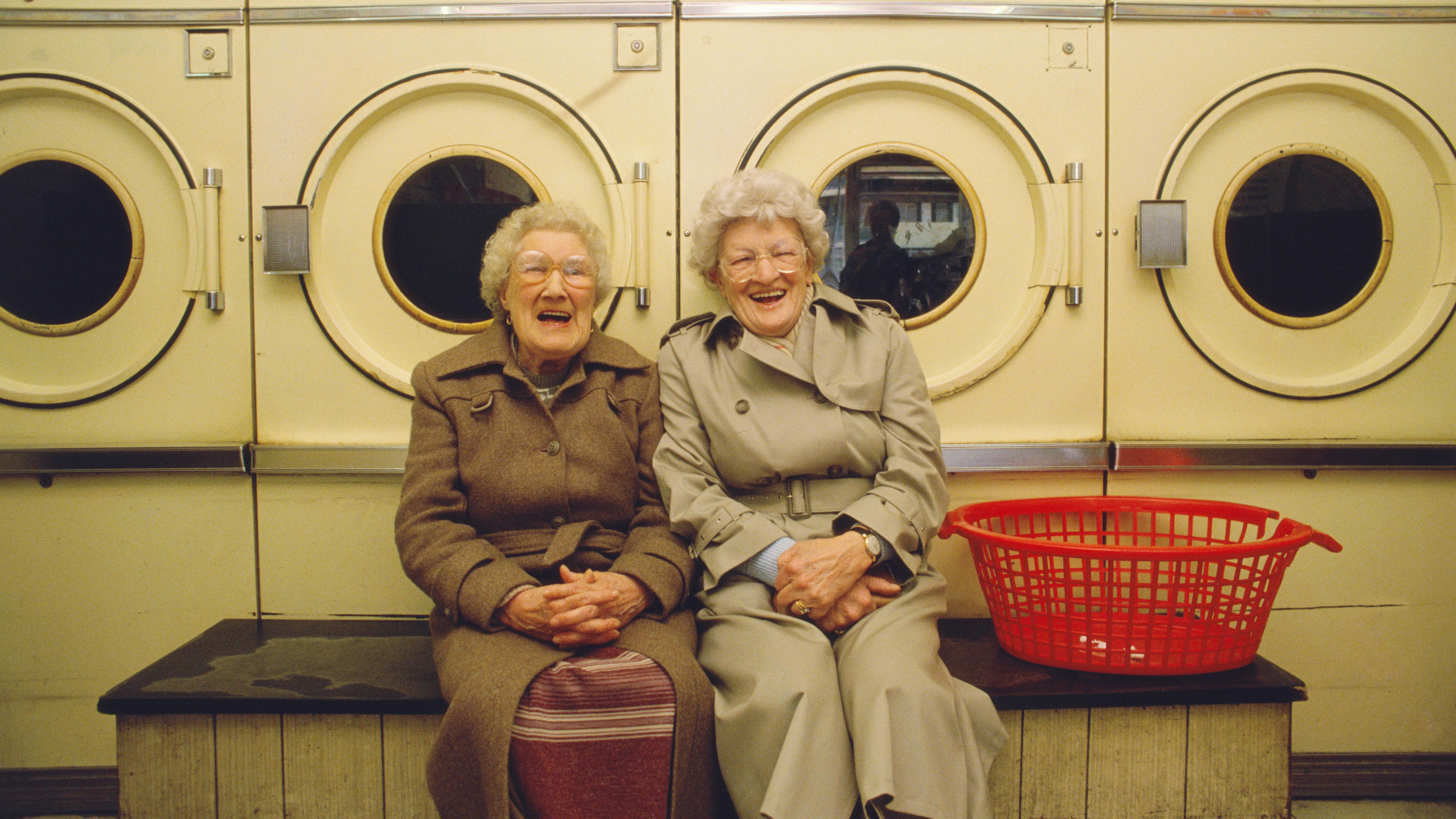 Zwei Seniorinnen sitzen im Waschsalon und lachen. 