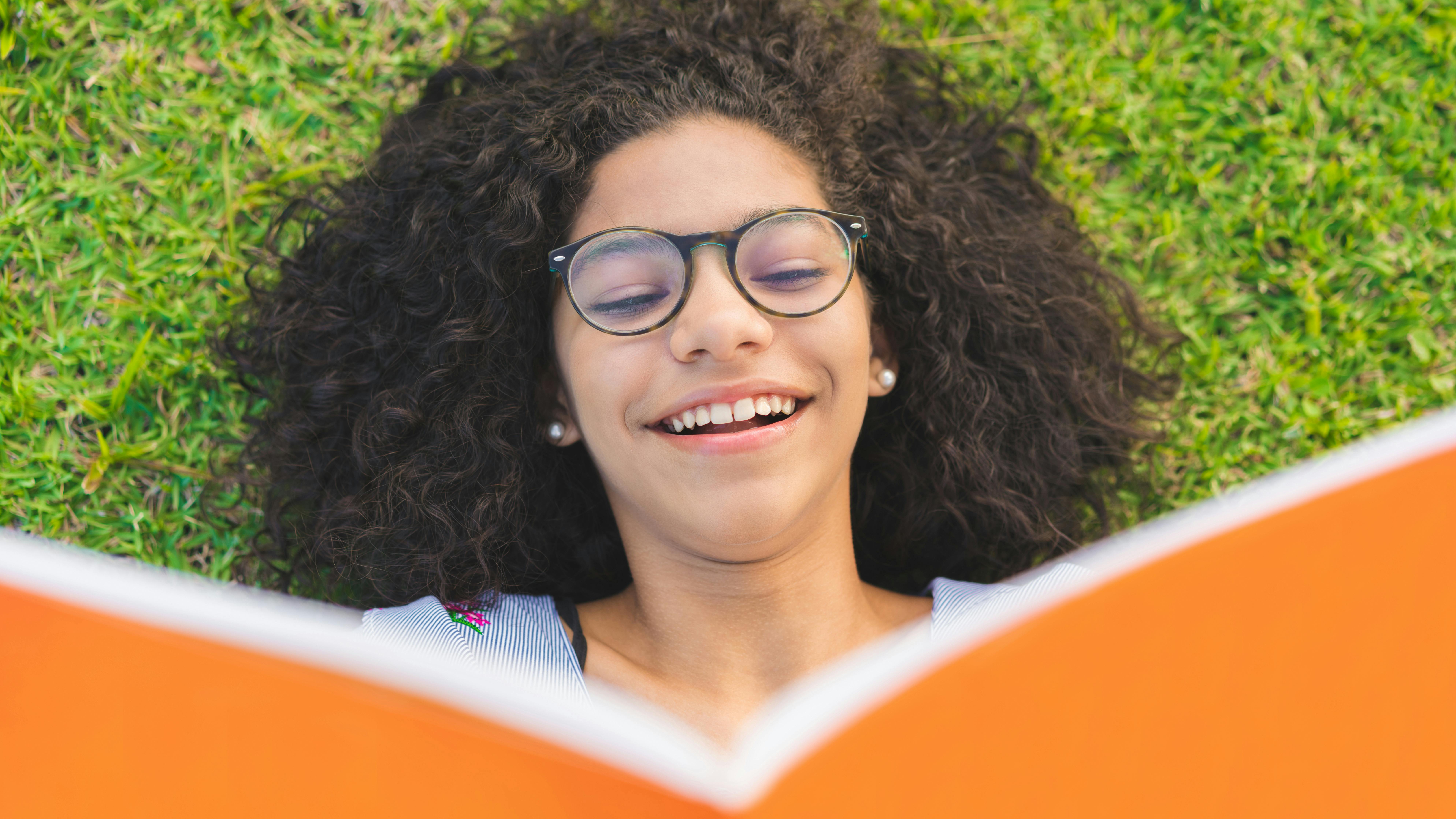 Eine junge Frau freut sich auf dem Rasen liegen, ein Buch lesend. 