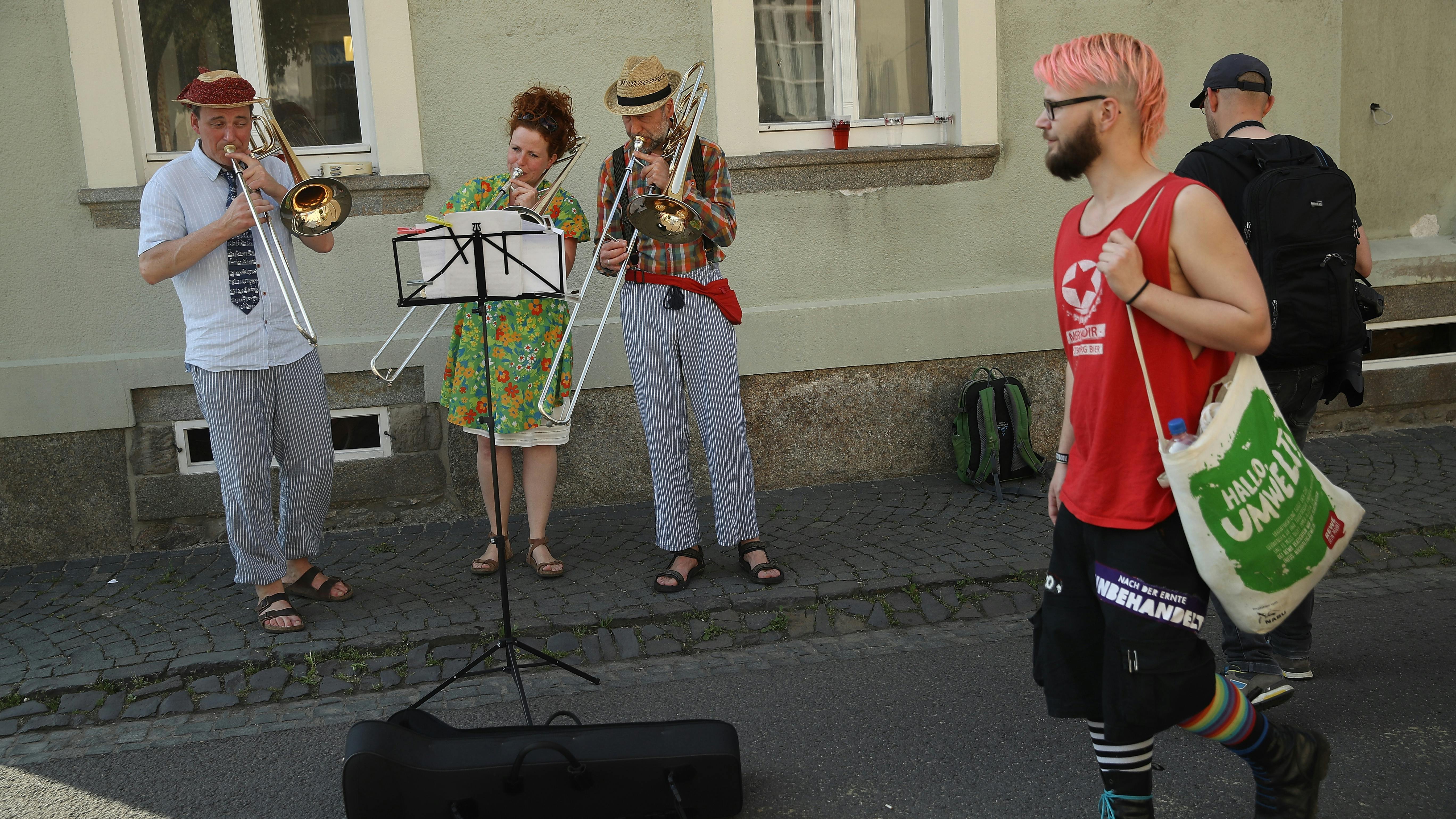 StraßmusikantInnen spielen auf offener Straße während eine Punkig- aussehende Person an ihnen vorbeischlendert. Die Menschen sind Teil eines zivil-gesellschaftlichen Bündnisses welches gegn ein Nazi-Rechts-Rock-Festival protestiert.