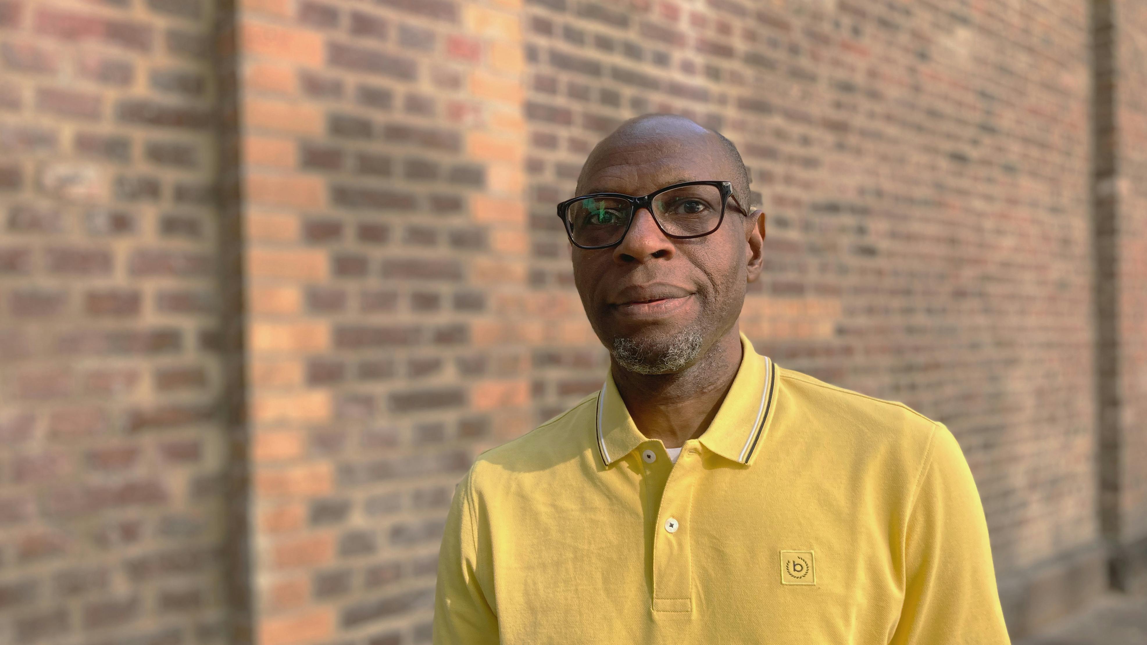 Ein schwarzer Mann mittleren Alters mit Brille vor einer Backsteinmauer