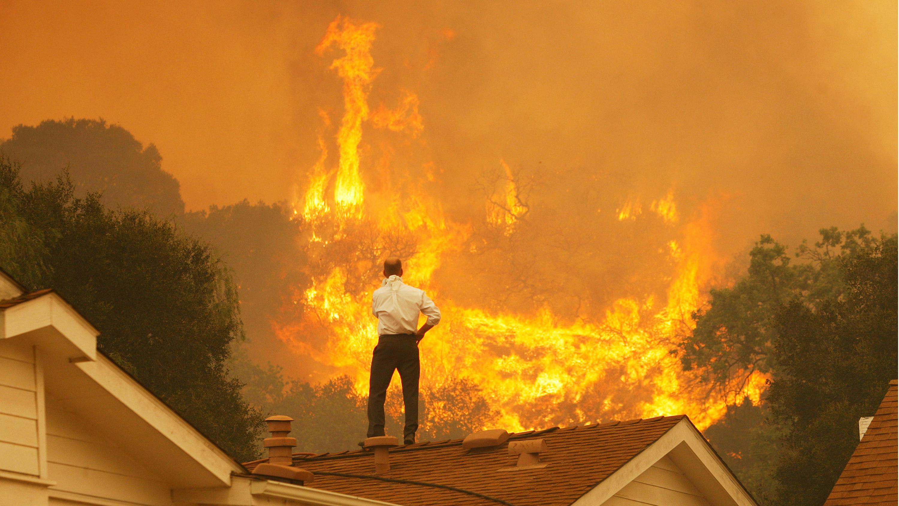 Ein brennendes Haus auf dessen Dach eine männlich gelesene Person steht.