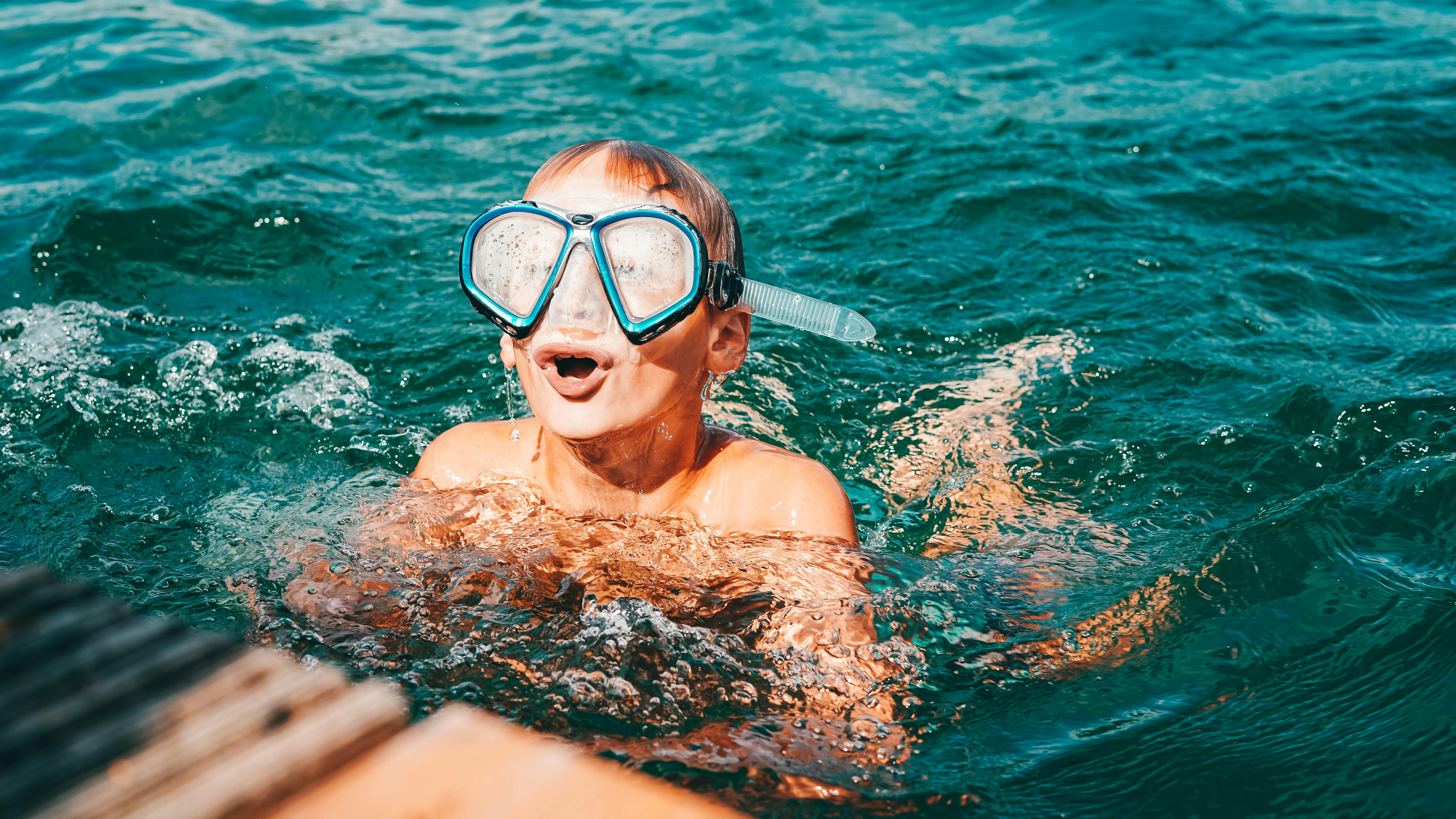 Ein Junge taucht mit einer Taucherbrille über den Augen aus dem Seewasser auf und holt tief Luft. 