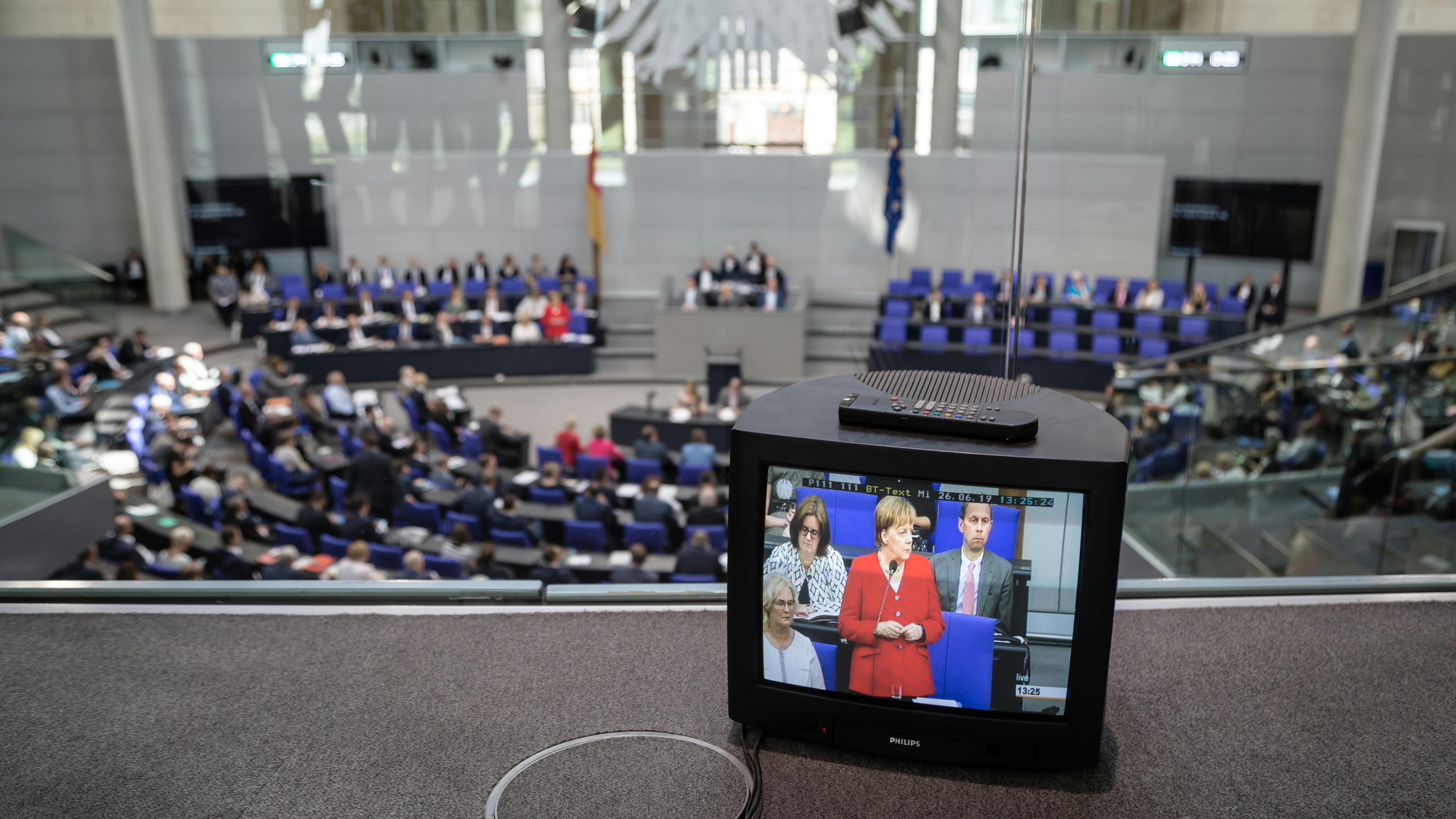 Ein Blick in den Plenarsaal des Bundestages. Im Vordergrund steht ein altmodischer Röhrenfernseher, auf dem Angela Merkel zu sehen ist. Im Hintergrund sitzen Abgeordnete auf den Rängen des Parlaments.