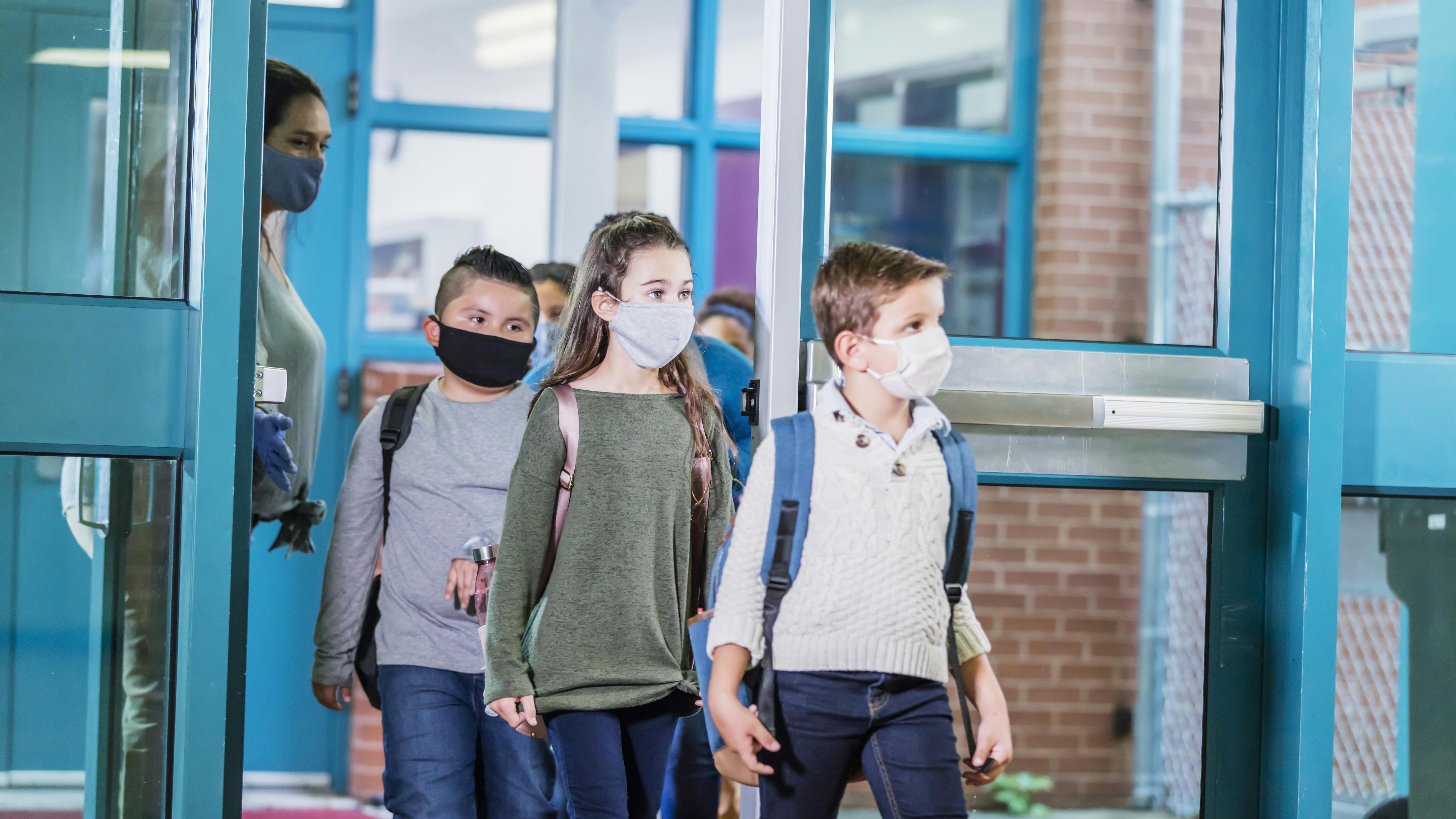 Vier Schüler:innen, mit Gesichtsmaske, gehen durch eine Tür aus der Schule heraus. Eine Lehrerin hält ihnen die Tür auf. 
