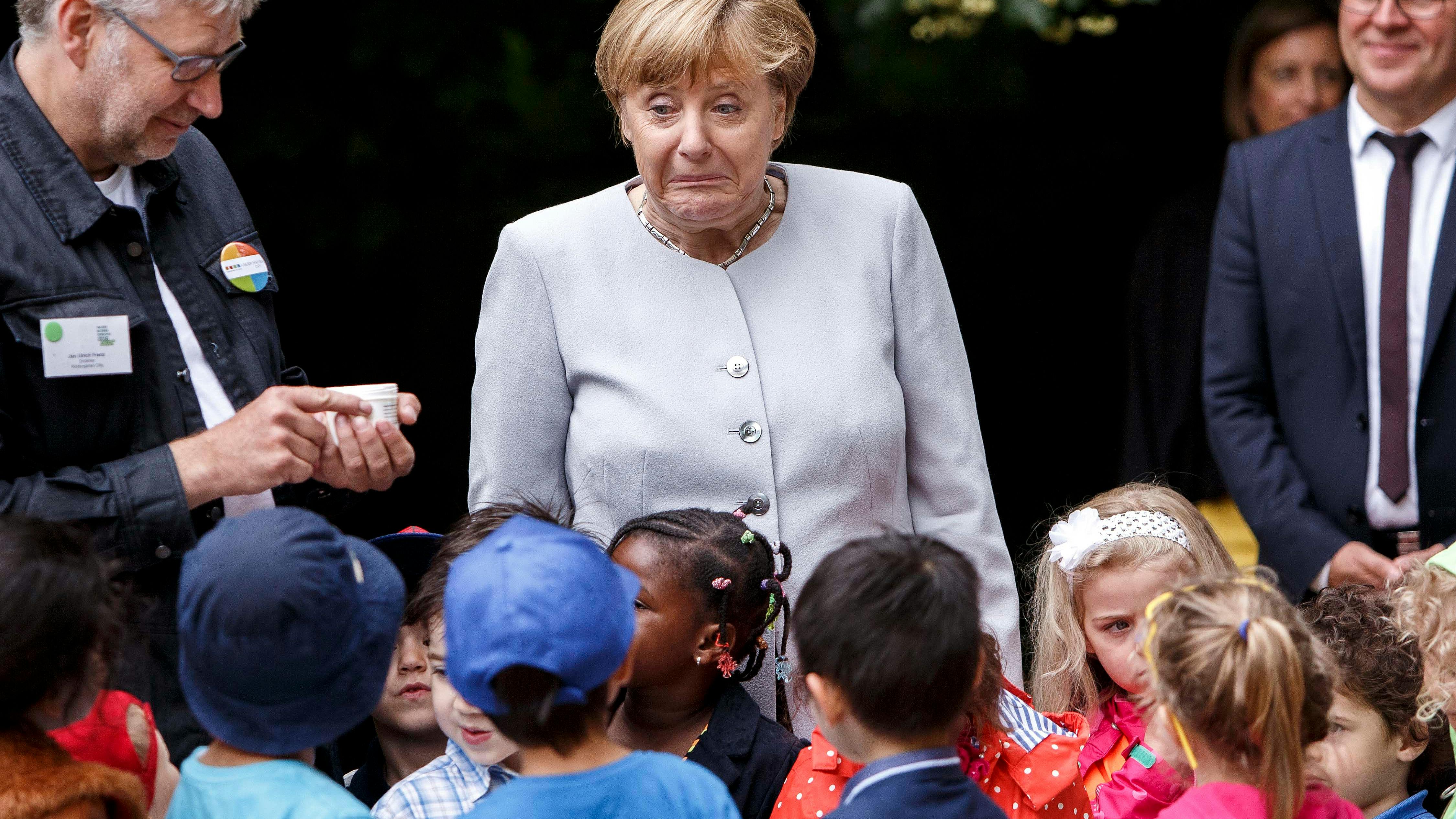 Angela Merkel steht mit wunderlich verzogenem Gesichtsaudruck in Mitten einer Gruppe Kinder.