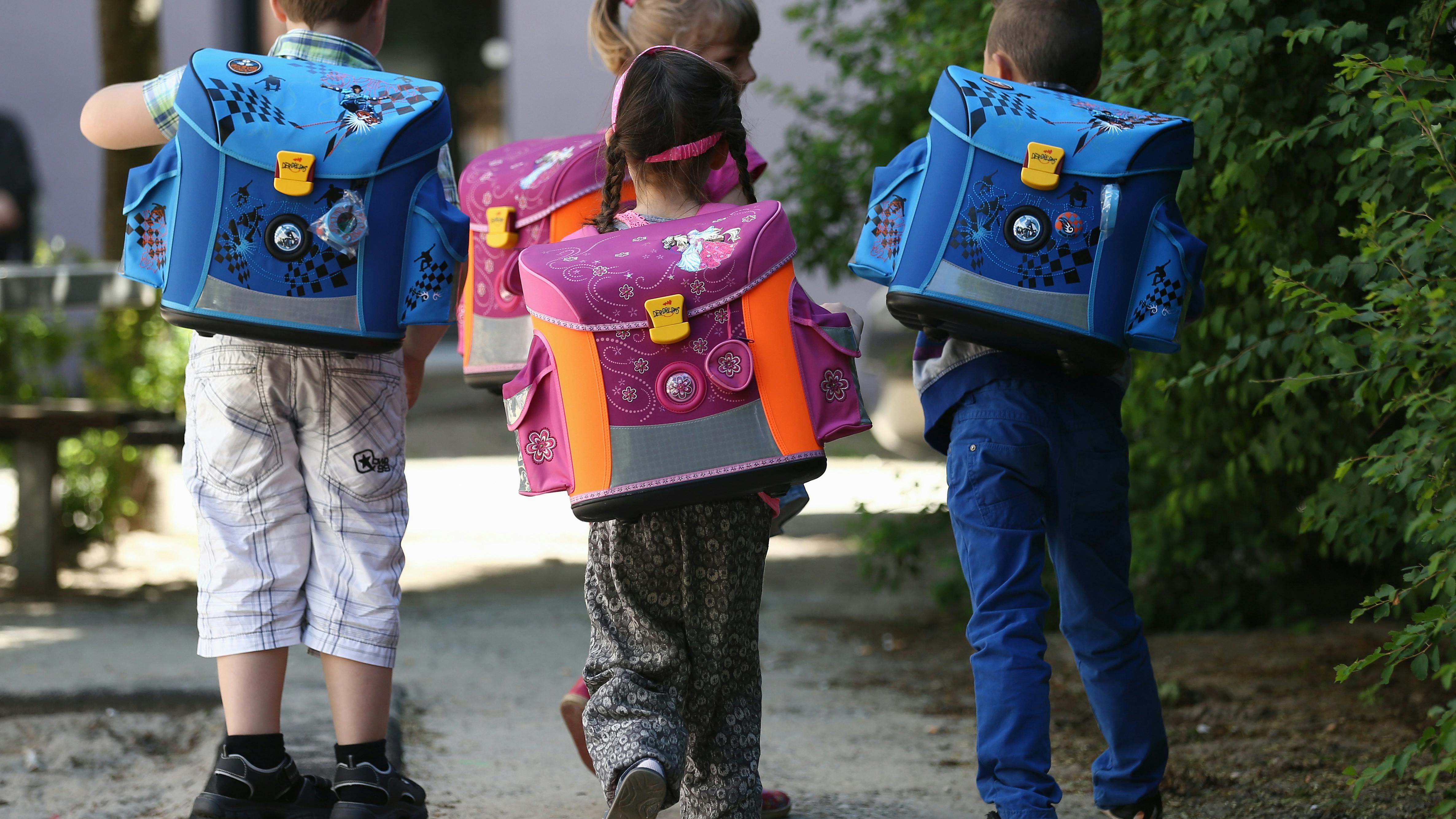 Vier Kinder laufen einen Weg entlang. Sie alle tragen Schulranzen auf dem Rücken, die Mädchen in rosa, die Jungen in blau.
