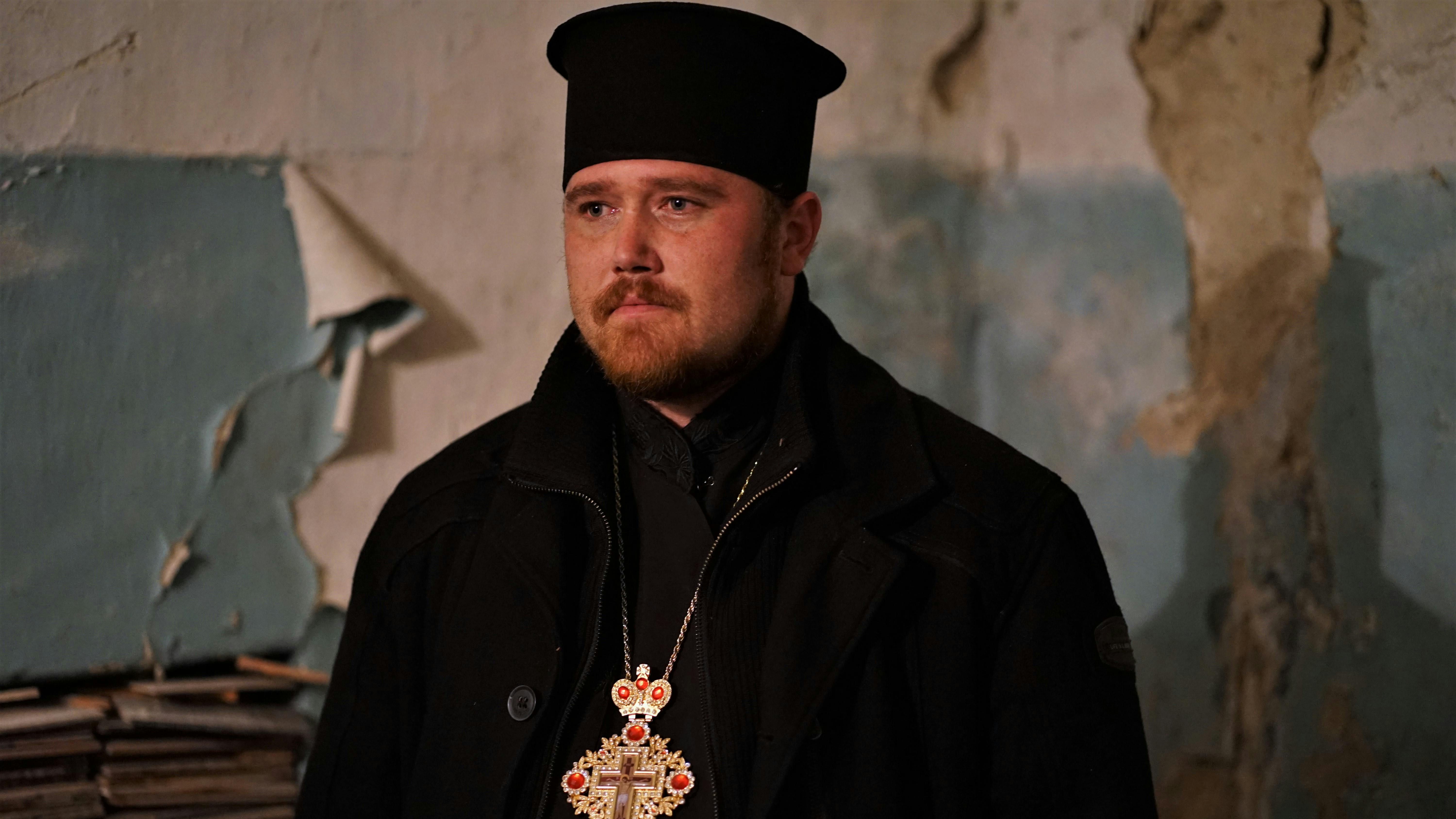 Dionissij Wassyljew trägt einen eine orthodoxen Popen und ein Kreuz um den Hals. Der Geistliche steht in dem Folterkeller in welchem er drei Tage verhört und gefoltert wurde. Sein Blick ist fest, doch wirkt er zerbrechlich.