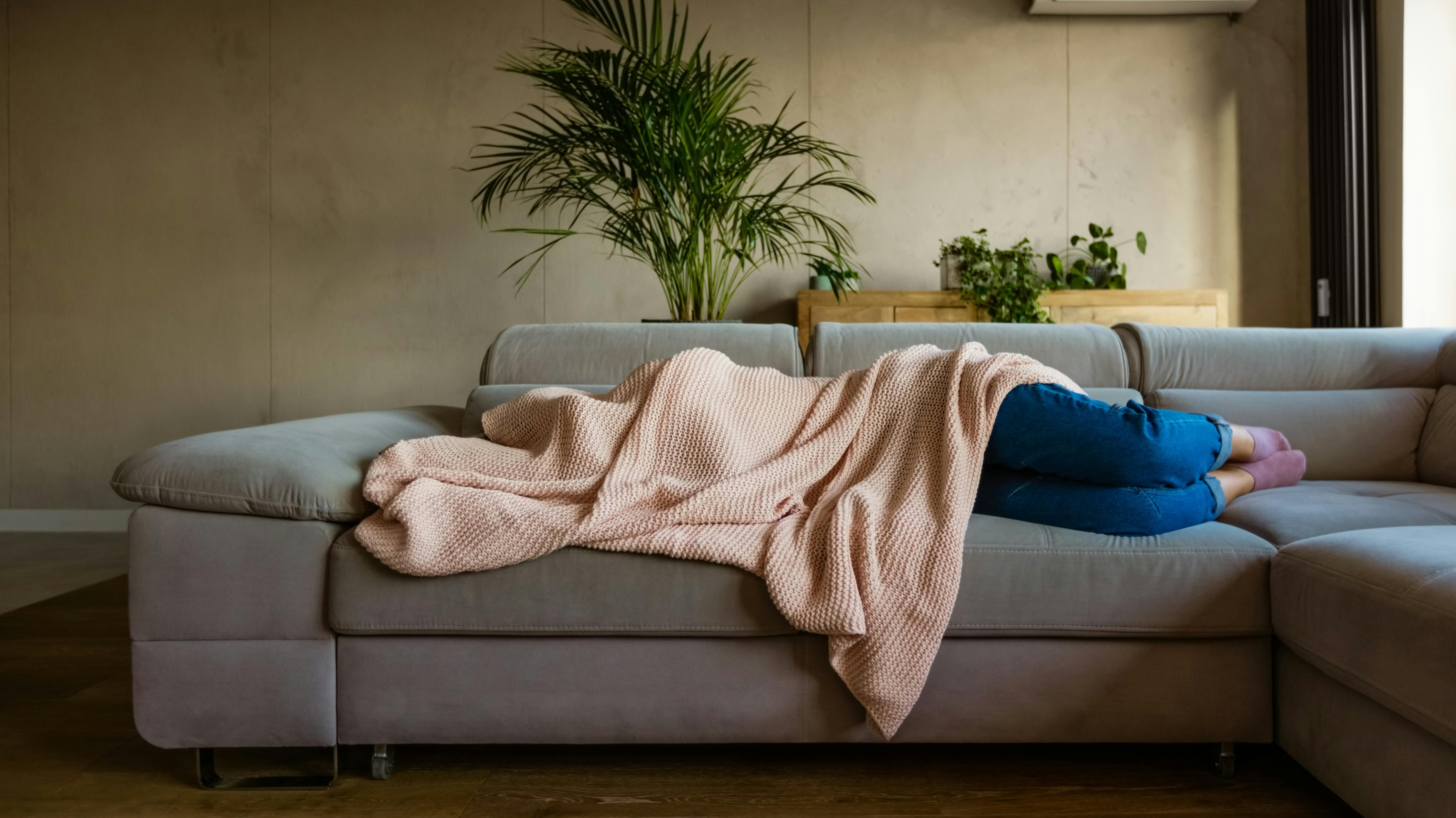 Eine Frau liegt auf dem Sofa und versteckt sich unter einer rosafarbenen Wolldecke.