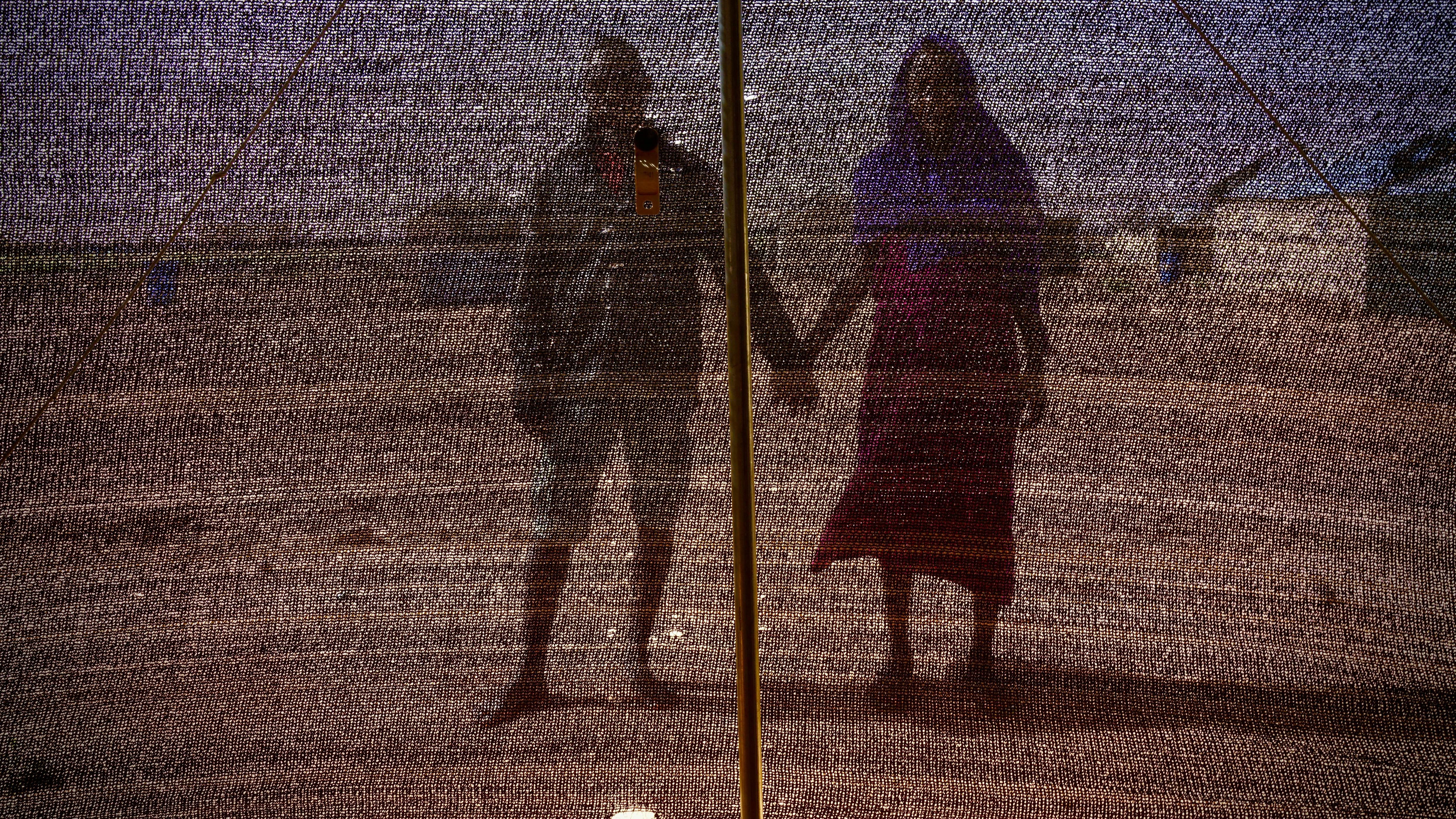 Zwei geflüchtete Menschen in Niger, scheinbar ein heterosexuelles Paar, halten Händchen, wir sehen ihre Silhouette durch ein schwarzes Maschennetz.