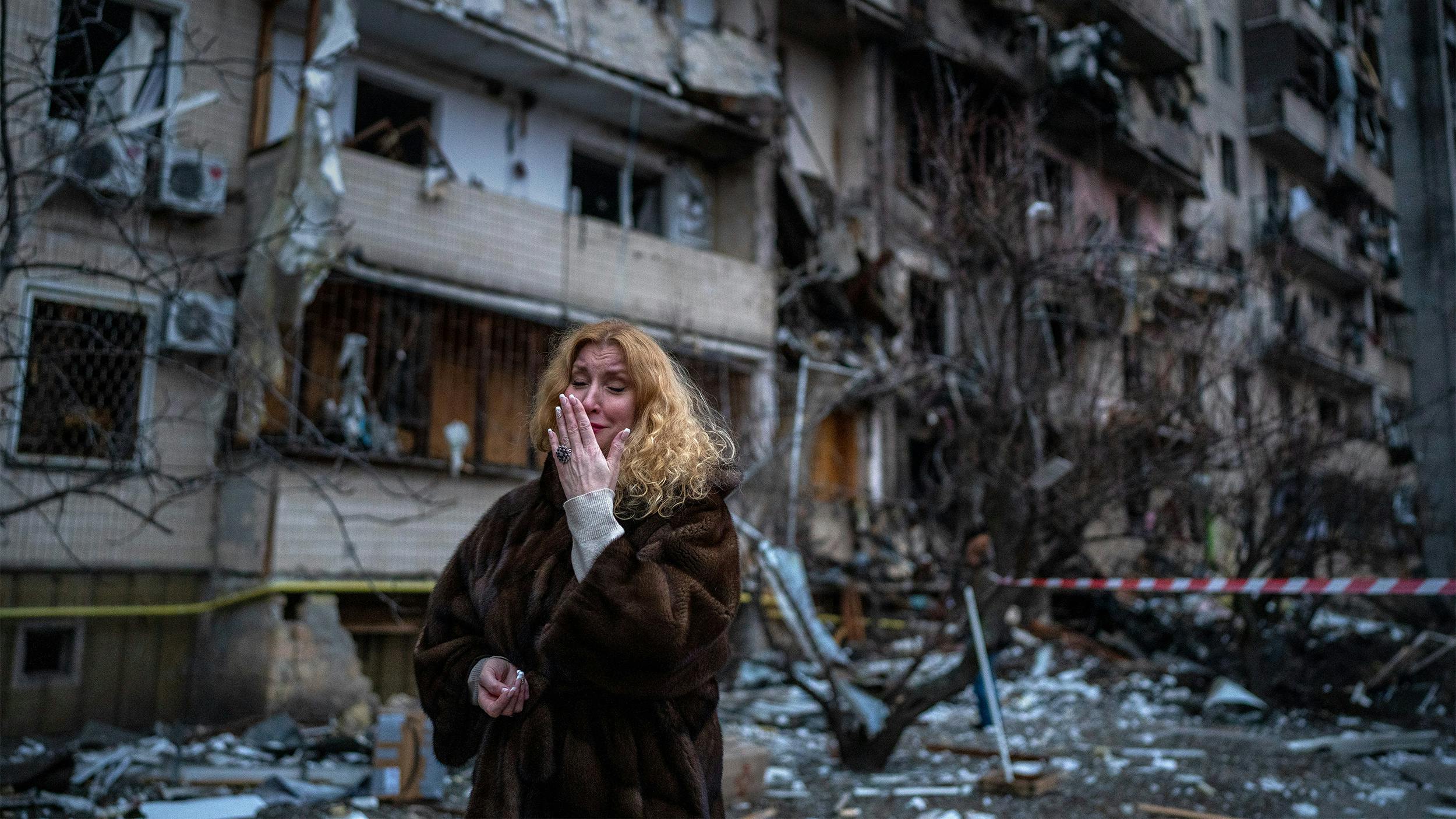 Eine Frau steht vor einem zerstörten Wohngebäude. Sie weint und hält die Hand vor ihr Gesicht. Sie trägt einen Pelzmantel.