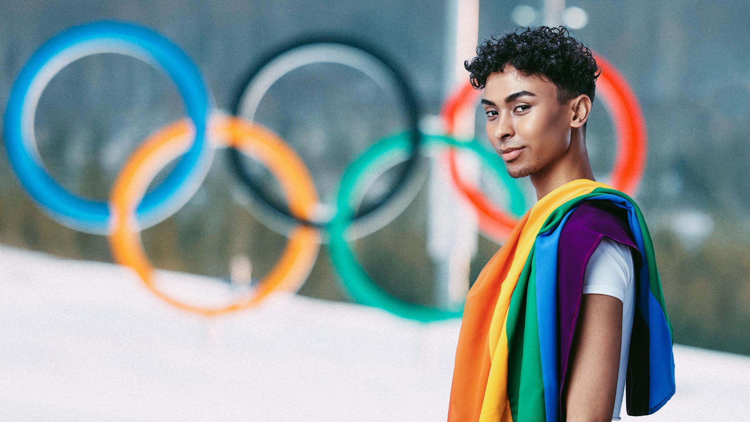 Ein junger Mann mit Regenbogenflagge über der Schulter blickt in die Kamera. Im Hintergrund sind die olympischen Ringe im Schnee zu sehen. 