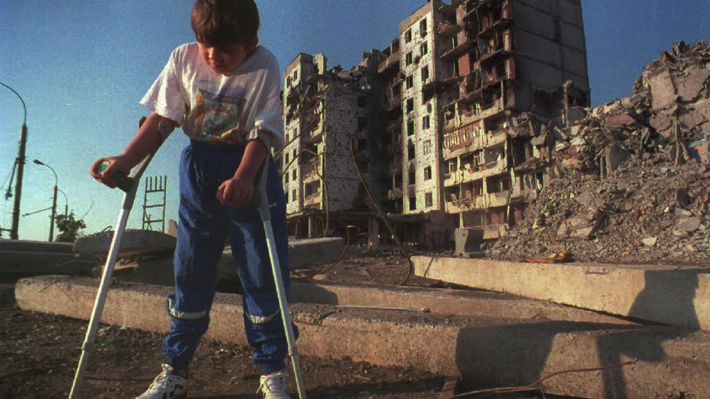 Ein Juge auf Krücken steht auf einer staubigen Straße, im Hintergrund ist ein zerbombtes Wohnhaus.