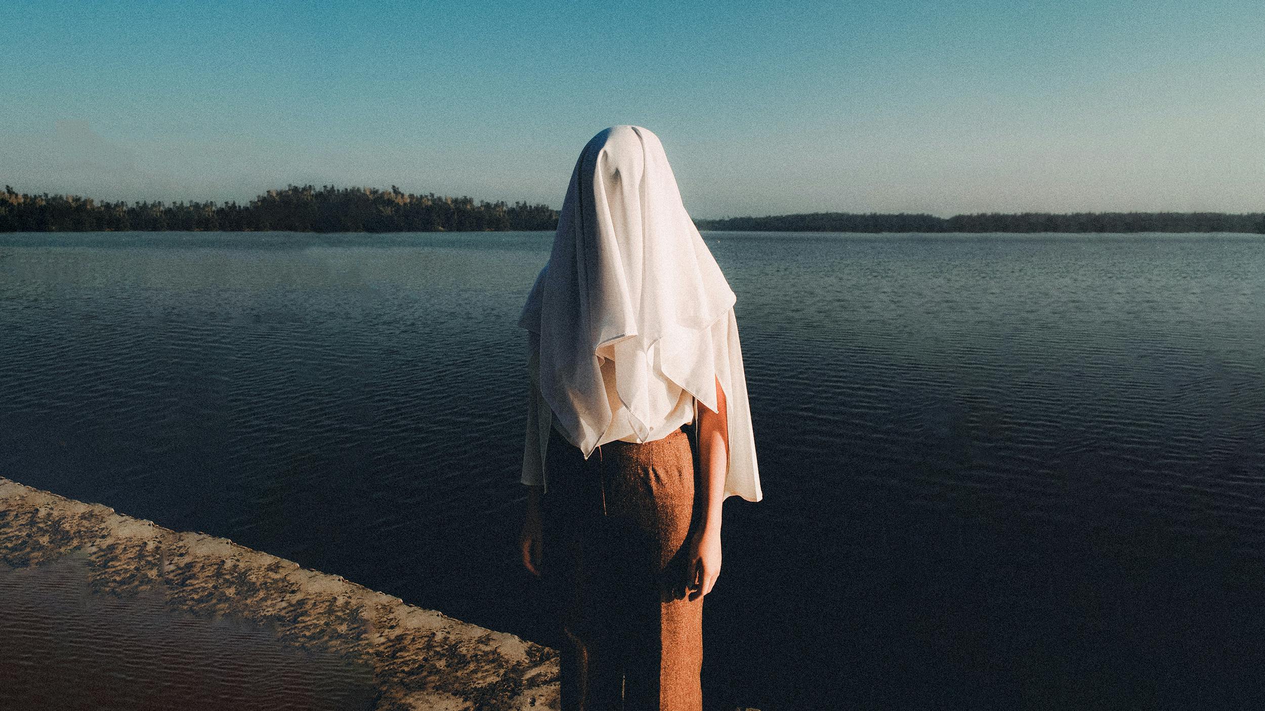 Eine Person steht am Strand eines Sees, über ihrem Kopf liegt ein Laken, dass den Kopf verdeckt.