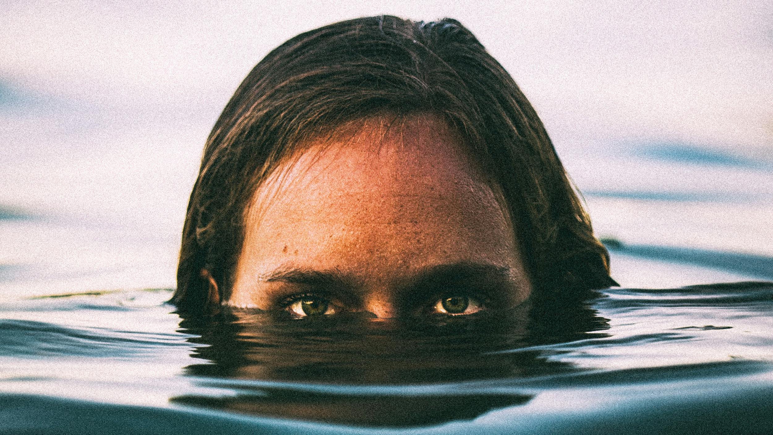 Man sieht einen Mann, wie er seien Kopf aus dem Wasser streckt und einen direkt anschaut. Sein Kopf guckt nur halb aus dem Wasser raus.