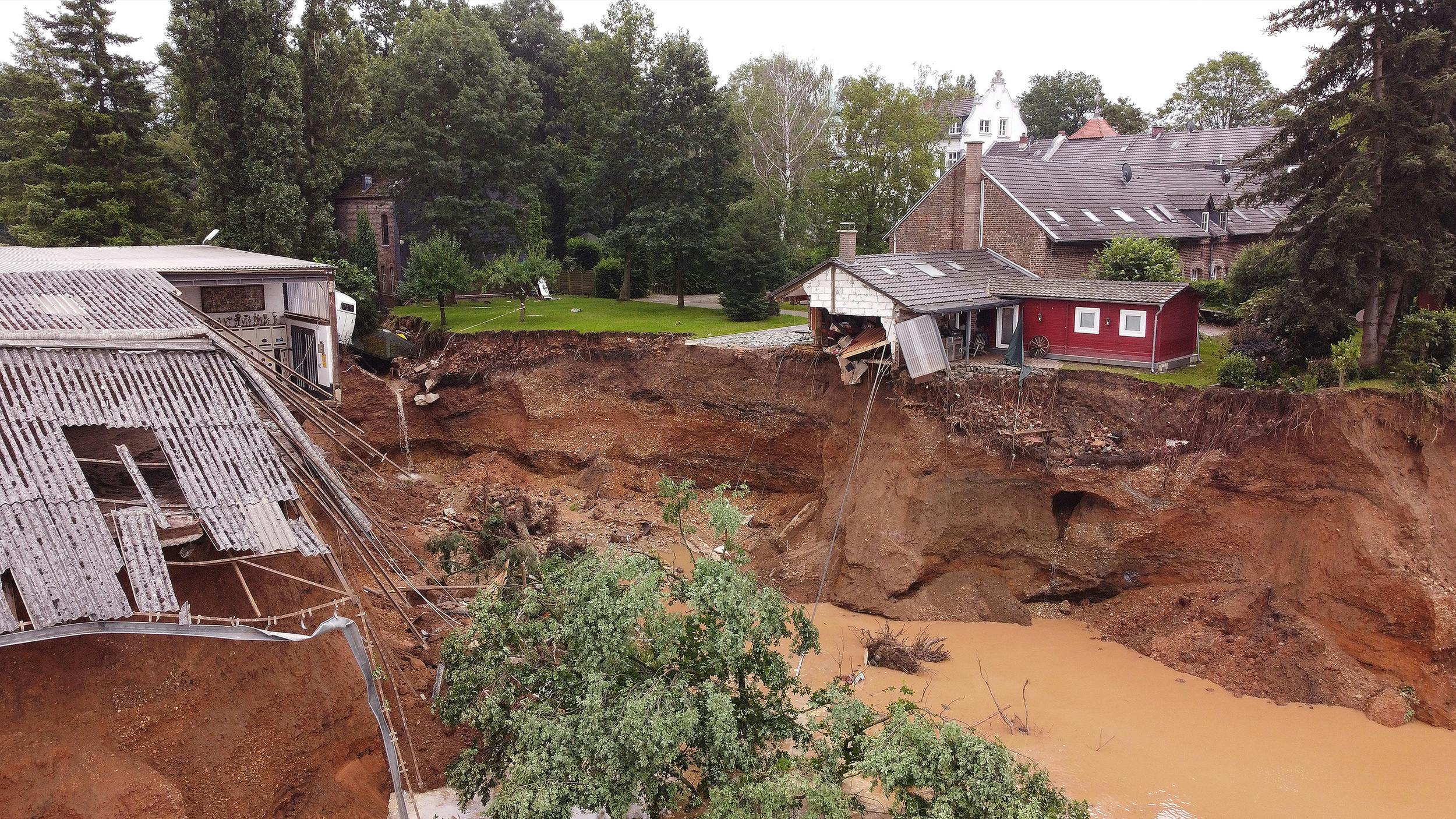 Ein Graben trennt zwei Gebäude voneinander. Beide weisen Beschädigungen auf. Im Graben fließt braunes Wasser. 