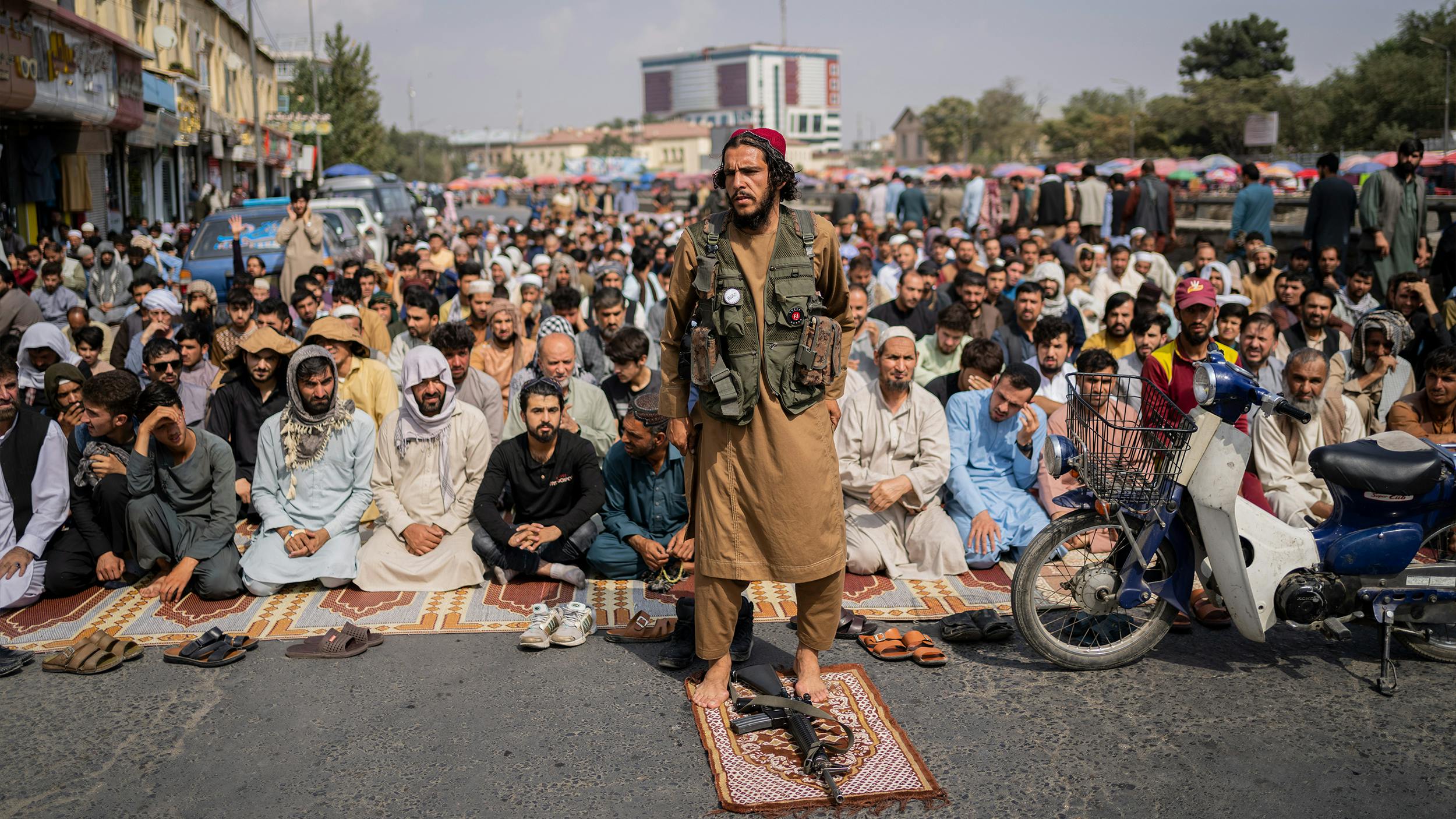 Ein Taliban-Kämpfer steht vor einer Gruppe betender Männer. Zu seinen Füßen liegt sein Gewehr auf einem roten Gebetsteppich