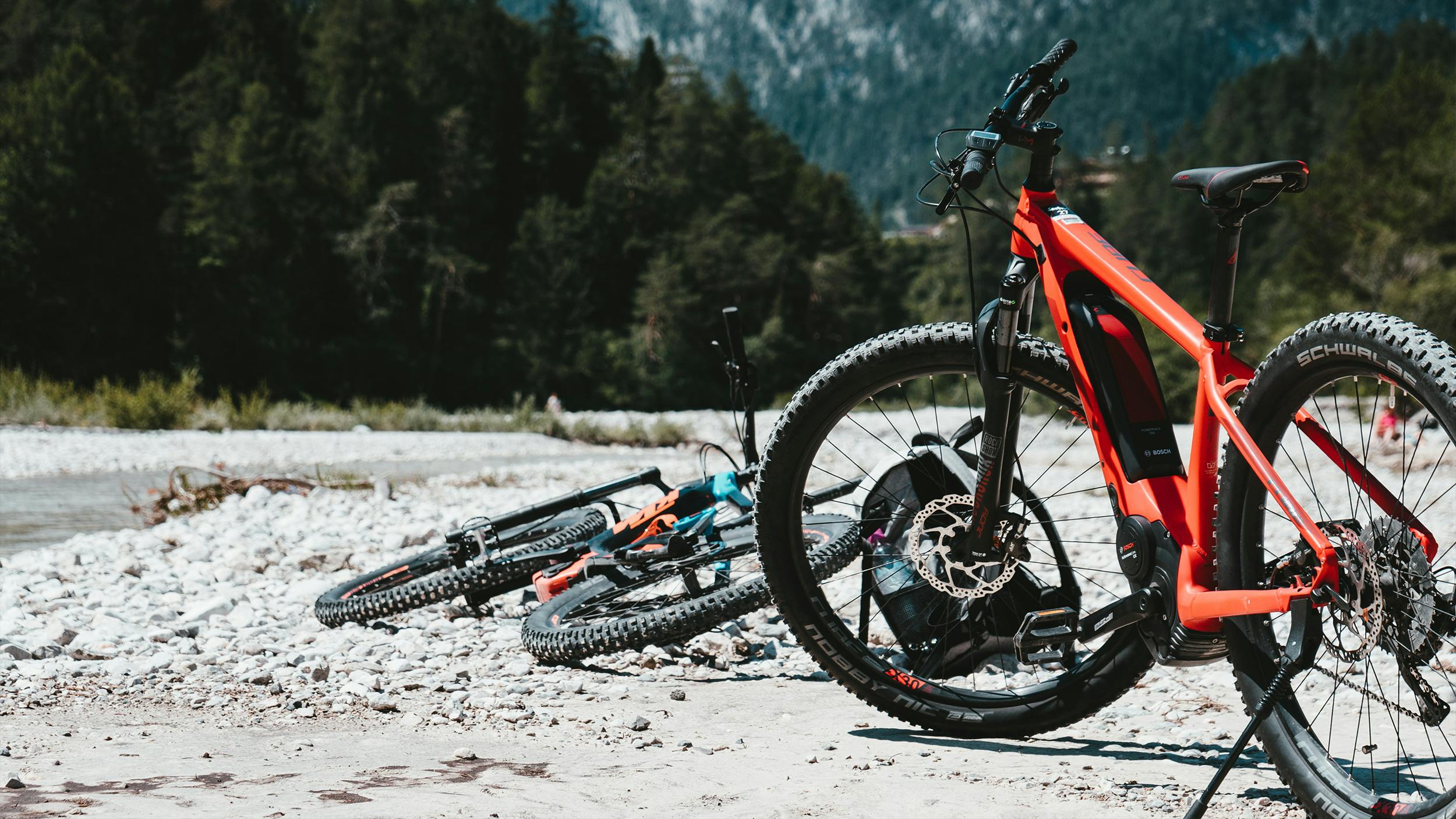 Ein Fahrrad steht im Vordergrund auf Schotter. Dahinter liegt ein zweites. Ein kleiner See lässt sich erahnen, dahinter sind bewachsene Berge.