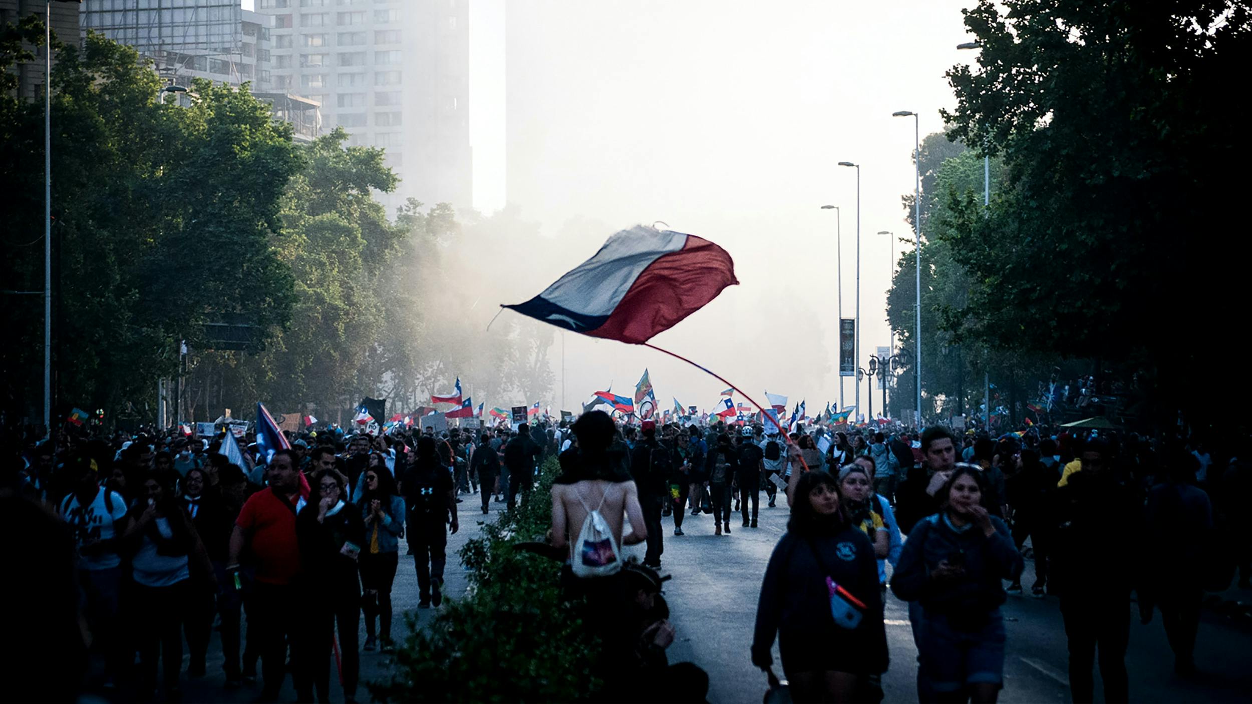 Auf einer von Bäumen gesäumten Straße sind viele Demonstrant:innen unterwegs, einige schwenken die chilenische Flagge. Im Hintergrund sind Hochhäuser zu erkennen, sie sind in Rauch oder Dampf gehüllt.